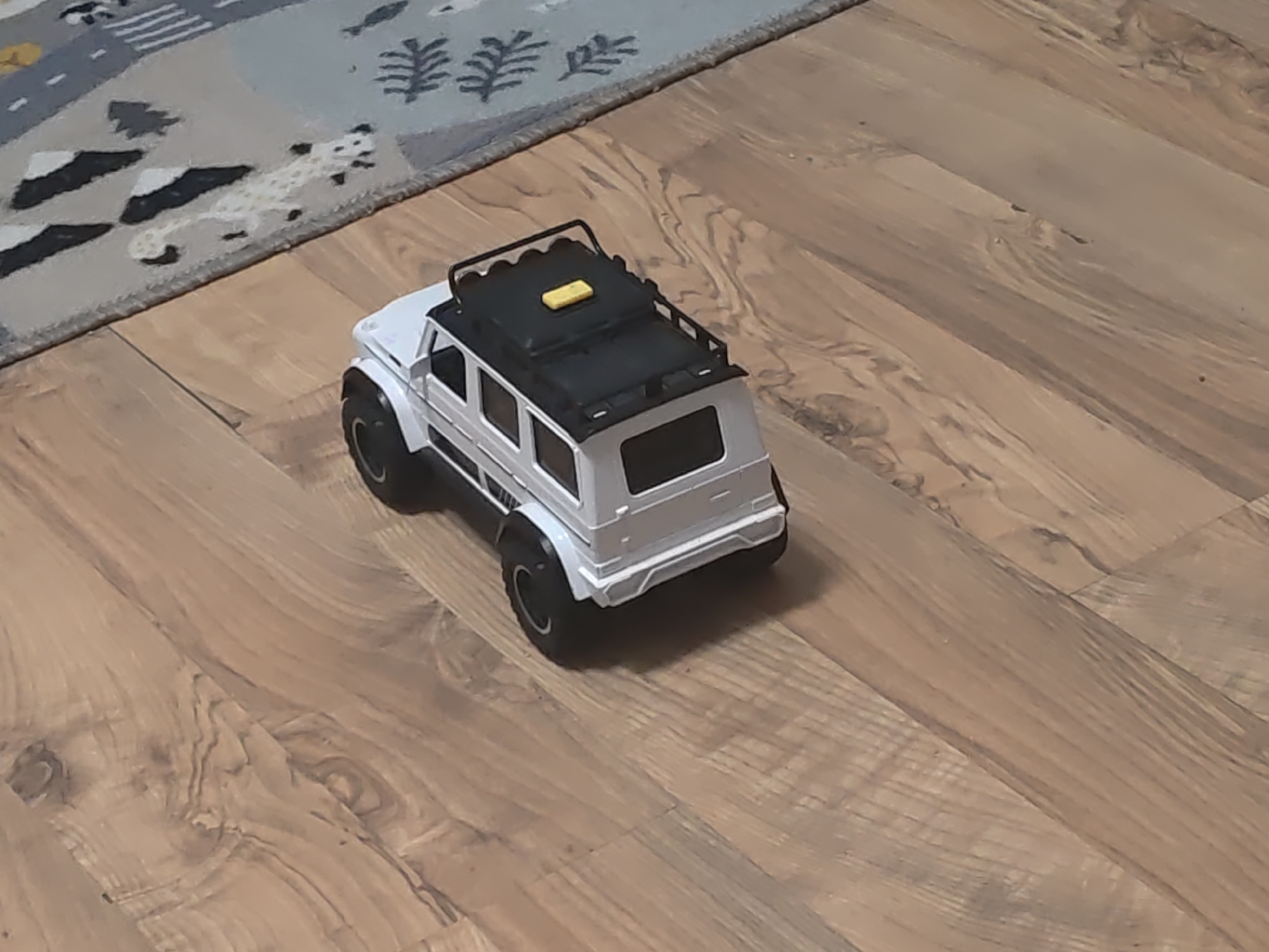 A white toy SUV with a black roof rack is positioned on a wooden floor. The toy car is facing away from the viewer, and its back end is slightly angled towards the left of the frame. The roof rack has a small yellow object on it. To the left of the toy car, a portion of a rug with a pattern of grey, white, and black shapes is visible. The wooden floor has a natural grain pattern and a slightly worn appearance. The scene is indoors and well-lit. The specific location in the city of Kurganinsk, Russia, cannot be determined from the visual information provided in the image.