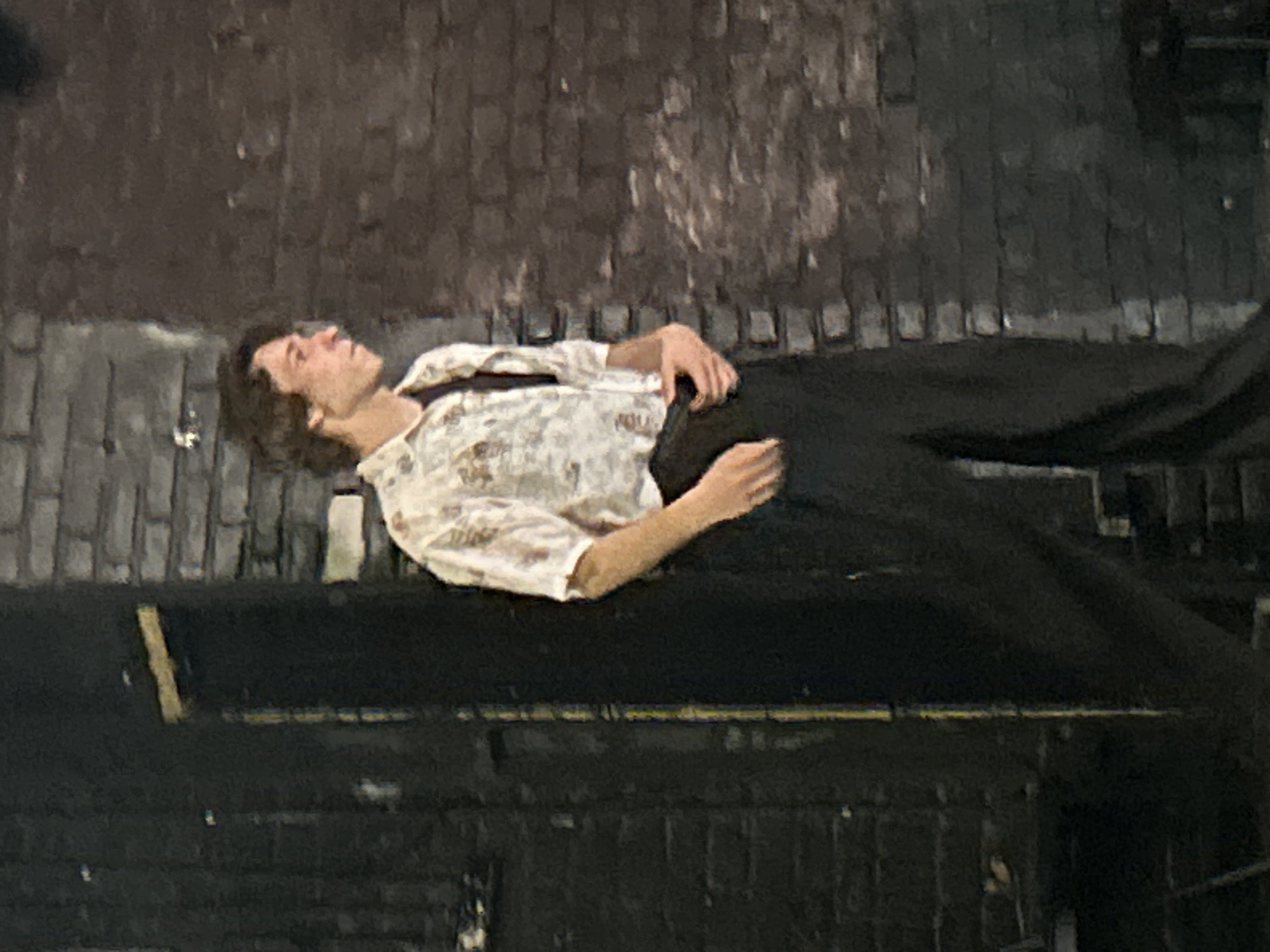 A young man with brown, wavy hair is standing in front of a brick wall. He is wearing a short-sleeved, patterned button-up shirt over a dark shirt, and dark pants. He is looking to his right, with his left hand on his pants. The background consists of a dark, textured surface and a brick wall. The lighting is dim.