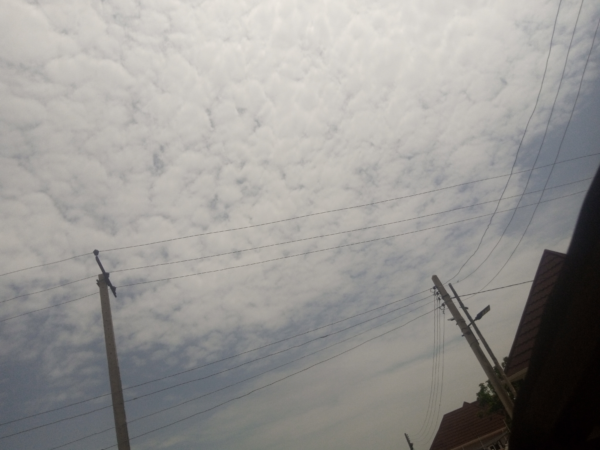 This is a low-angle, outdoor photograph taken in Gura Top, Nigeria, featuring a cloudy sky with a pattern of cumulus clouds. Several utility poles with overhead wires are visible, two of which are prominent in the foreground. The pole on the left is a tall, light-colored concrete pole with wires extending horizontally. To the right, another similar pole is angled, supporting multiple wires. In the far right of the image, a dark shadow, possibly from a person or object, obscures part of the scene, and a portion of a tiled roofline is also visible. The overall impression is of a street scene on an overcast day.
