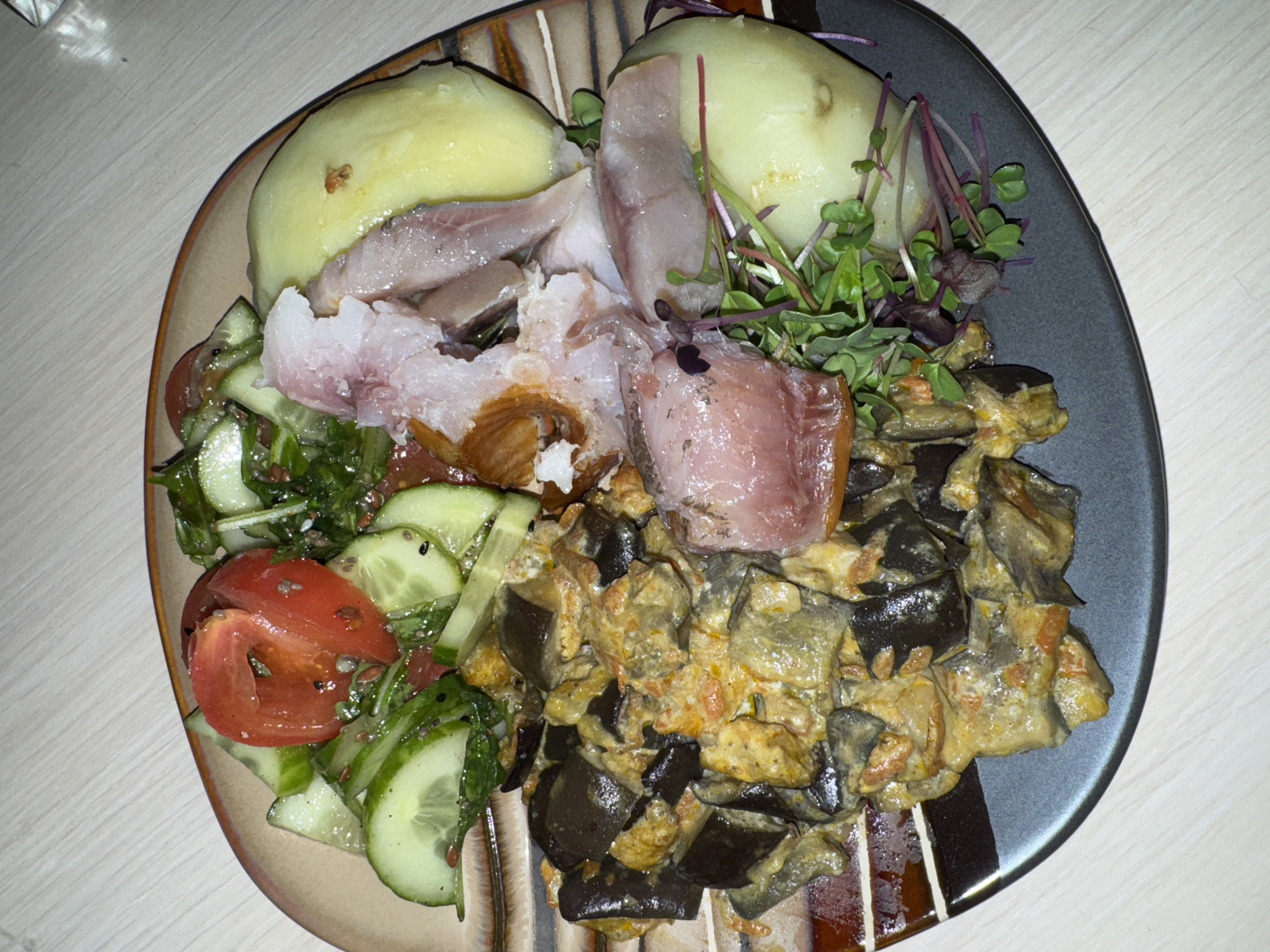 A plate of food is presented on a table. The plate contains boiled potatoes, pieces of smoked fish, a salad with sliced cucumbers and tomatoes, microgreens, and a dish made with eggplant. The image does not reveal any information about the location, the city of Chaikovsky, Russia, people, actions, interactions, or environment beyond what is directly visible in the plate of food.