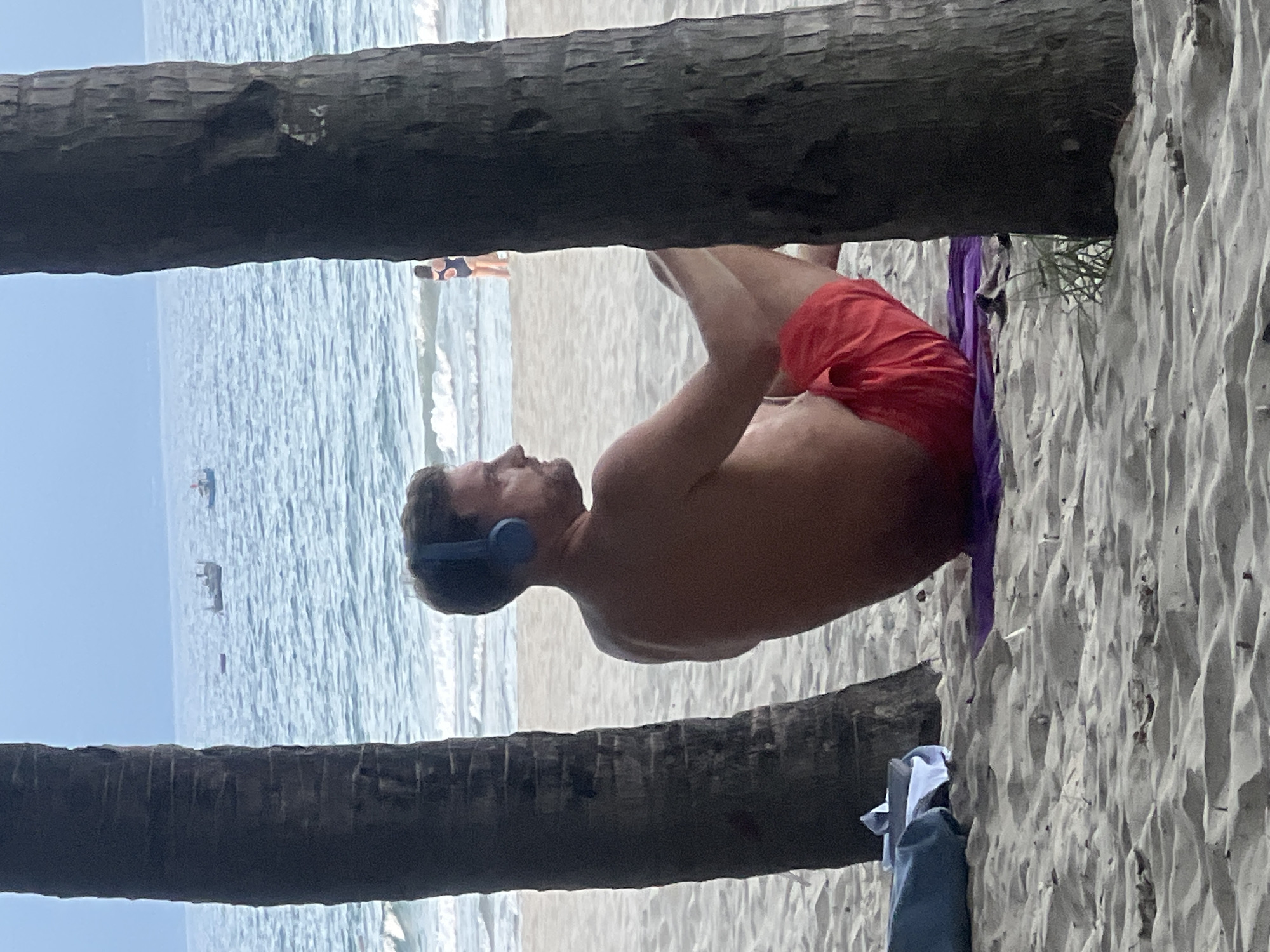 The image shows a man wearing blue headphones and red swim trunks sitting on a purple towel on a sandy beach. He is facing away from the camera, looking out at the ocean. Two palm trees frame the scene, with the ocean visible in the background. There are a few boats on the water and another person in the distance wearing a dark swimsuit with a watermelon pattern. The sky is overcast. The location is likely a beach in Da Nang, Vietnam.