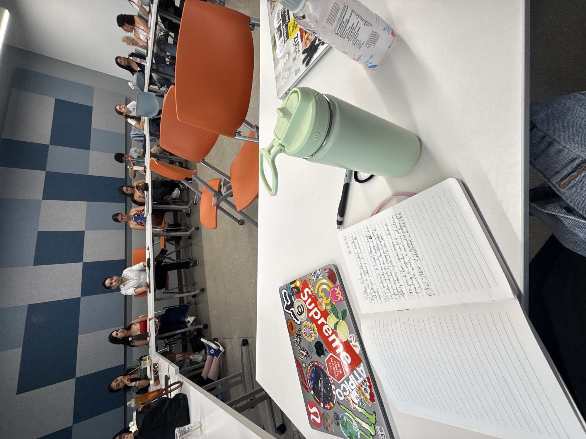 An eye-level shot captures a classroom or lecture hall scene, with the perspective centered on a white table in the foreground. Several students are seated at tables arranged in rows, facing towards the front (left side of the image). Most students appear to be young adults, dressed in casual attire. Some are looking forward, others are looking down at their tables or devices. The room features a distinctive wall design with large blue and grey checkered panels.
In the foreground, on the white table, a light mint green Lululemon water bottle with a handle stands prominently. Next to it is an open spiral-bound notebook with handwritten notes and ruled blank pages. A black pen rests near the notebook. To the left of the notebook is a silver laptop adorned with numerous colorful stickers, including a large red "Supreme" logo, a butterfly, a "Monster Energy" logo, and other varied designs. A small, clear plastic bottle with a white label is also visible at the top right of the table.
Visible text includes:
* On the laptop stickers: "Supreme", "ATIPICO", "ZU", "NO ME HABLES, FÁCIL ME DISTRAIGO".
* In the notebook: "16.04.26" (date), "Filipe Caldario" (name), and handwritten notes in what appears to be Spanish or Portuguese, with phrases like "Politica de Clientes", "rigida", "Detena fundamental", and "mundos corporales".
The setting suggests an educational environment during what appears to be a class or study session in Mexico City, Mexico. The lighting is bright and even, indicating an indoor, likely daytime, setting.