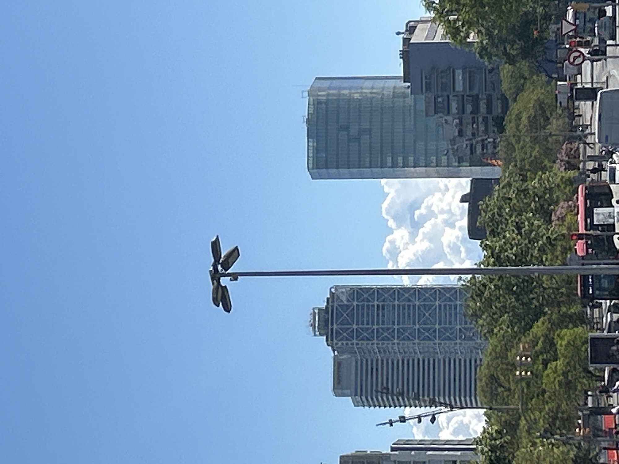 The image shows a street scene in Barcelona, Spain. In the foreground is a street lamp. Behind it are two prominent buildings: Torre Glòries, and behind it, the Hotel Arts Barcelona. There are clouds in the sky, and trees and traffic are visible on the right side of the image.