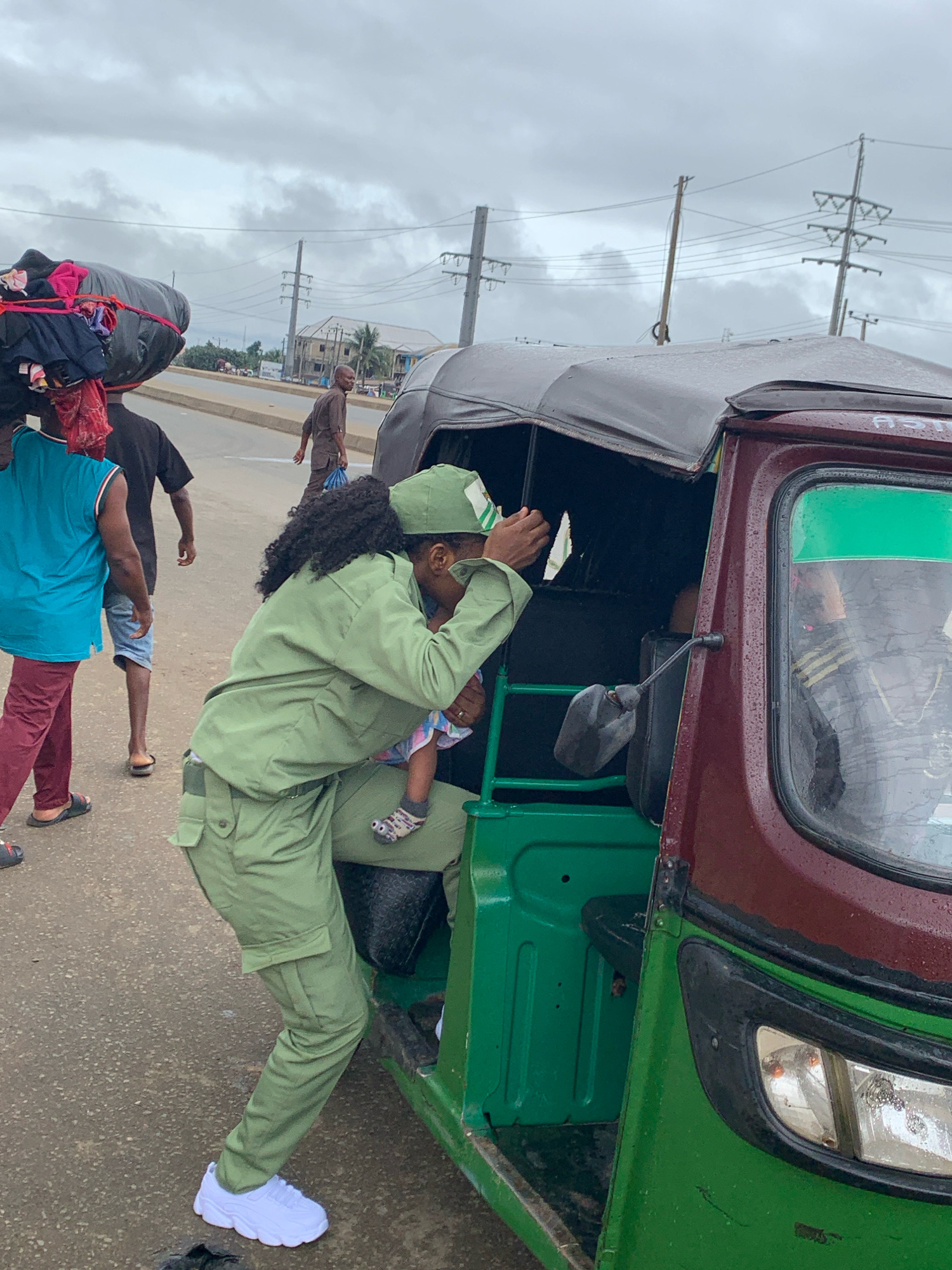 In Umuode, Nigeria, under a cloudy sky, a woman wearing a green uniform and cap is helping a child into a three-wheeled vehicle, commonly known as a Keke. The woman is bending down, holding the child who is wearing a striped sock on one foot. Her green uniform consists of a jacket and cargo pants, and she is wearing white sneakers. Another person, wearing a blue sleeveless shirt and maroon pants, stands to the left, with a large bundle of clothes and personal items strapped to their head. Behind this individual, a person in a dark shirt and blue shorts walks away from the Keke. The Keke itself is predominantly green with a maroon-colored top and appears to have a canvas cover. The background shows a paved road with utility poles and power lines overhead, indicative of a typical urban or semi-urban setting. The road surface shows signs of moisture, suggesting recent rain.