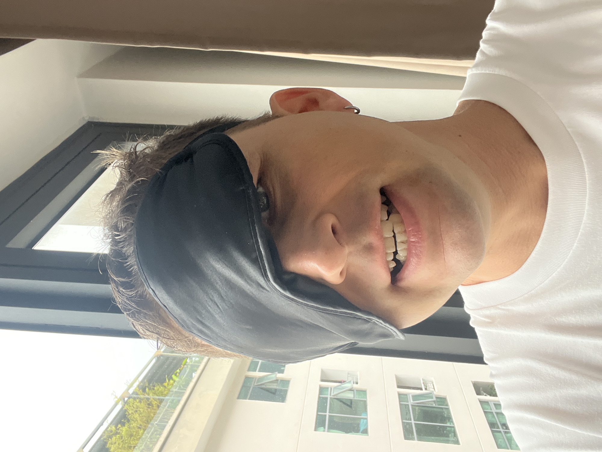 A person wearing a black eye mask is smiling. The person is wearing a white shirt. Behind them are windows with green plants visible outside, suggesting a balcony. The windows and architecture appear to be in the style of buildings found in Bac Nha Trang Ward, Vietnam. The person is indoors.