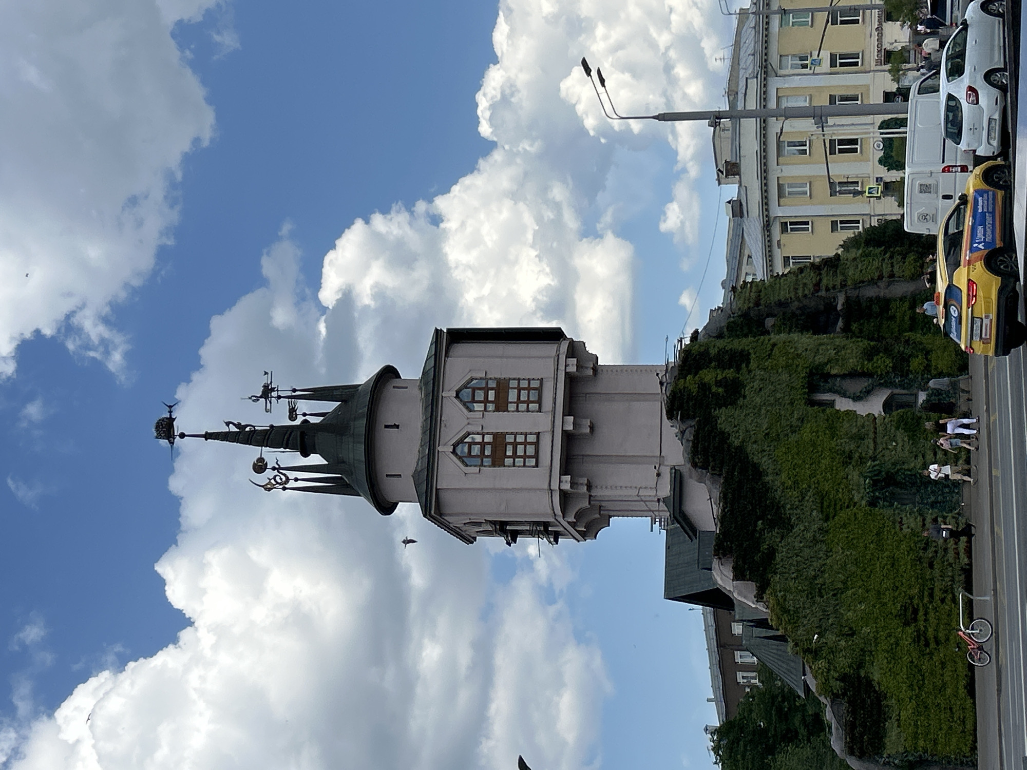 This image captures a vibrant urban scene in Moscow, Russia, dominated by a distinctive tower against a bright, cloudy sky. The photograph is taken during the daytime, under a clear blue sky dotted with large, fluffy white clouds, suggesting sunny weather.
The most prominent subject is a light-colored, possibly pale pink or beige, tower with a dark green, conical roof. Its spire is adorned with multiple ornate finials and weather vanes, one of which appears to be a bird-like figure. The tower's main body features several rectangular windows with dark frames, arranged in a distinctive pattern.
In the middle ground, a sloped structure or wall is entirely covered in lush green foliage, creating a natural barrier that separates the foreground from the street scene beyond. Above this greenery, several buildings are visible. One notable building has a light yellow facade with numerous windows and balconies.
On the street, a white commercial van and a bright yellow taxi are parked or moving. The taxi has prominent blue and orange decals. Several people are visible walking on the sidewalk near the vehicles and the green embankment. A red bicycle is also parked near the curb. Streetlights and utility poles rise towards the sky.
Visible text on the yellow taxi reads "ЯНДЕКС ТАКСИ" (Yandex Taxi) and "УДИВИТЕЛЬНЫЙ МИР НАЧИНАЕТСЯ" (The amazing world begins). Faintly visible on the yellow building in the background is the text "ШОКОЛАДНИЦА" (Shokoladnitsa), indicating a popular coffee shop chain. The overall scene depicts a typical, bustling urban environment in Moscow.
