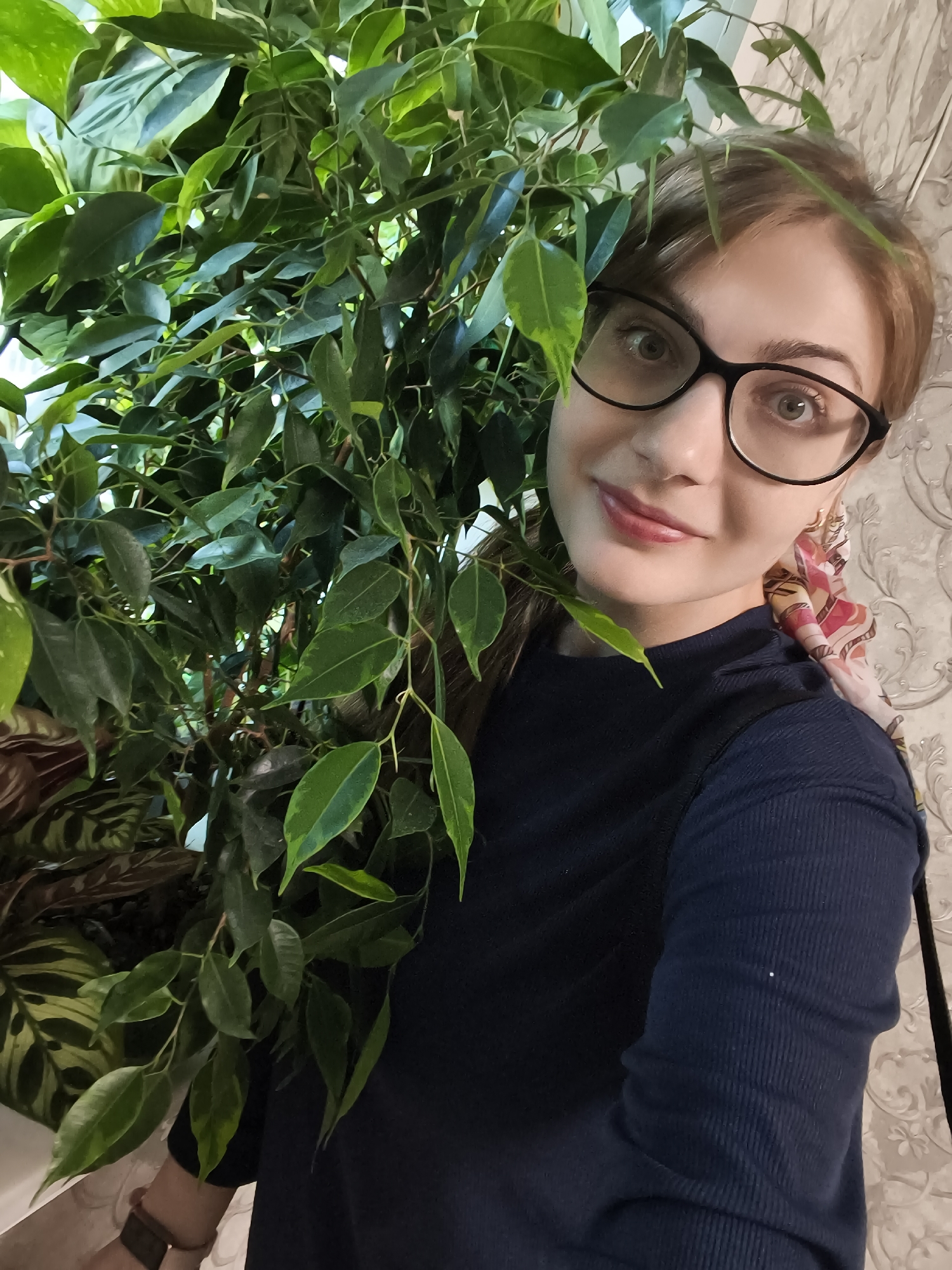 In Novaya Usman, Russia, a young woman with short blonde hair and glasses is taking a selfie in front of a large indoor plant. She is wearing a dark blue long-sleeved shirt and a colorful scarf around her neck. The plant has many green leaves and branches, some of which obscure parts of her face and body. The background is a light-colored, textured wall. The lighting suggests an indoor environment, possibly near a window.