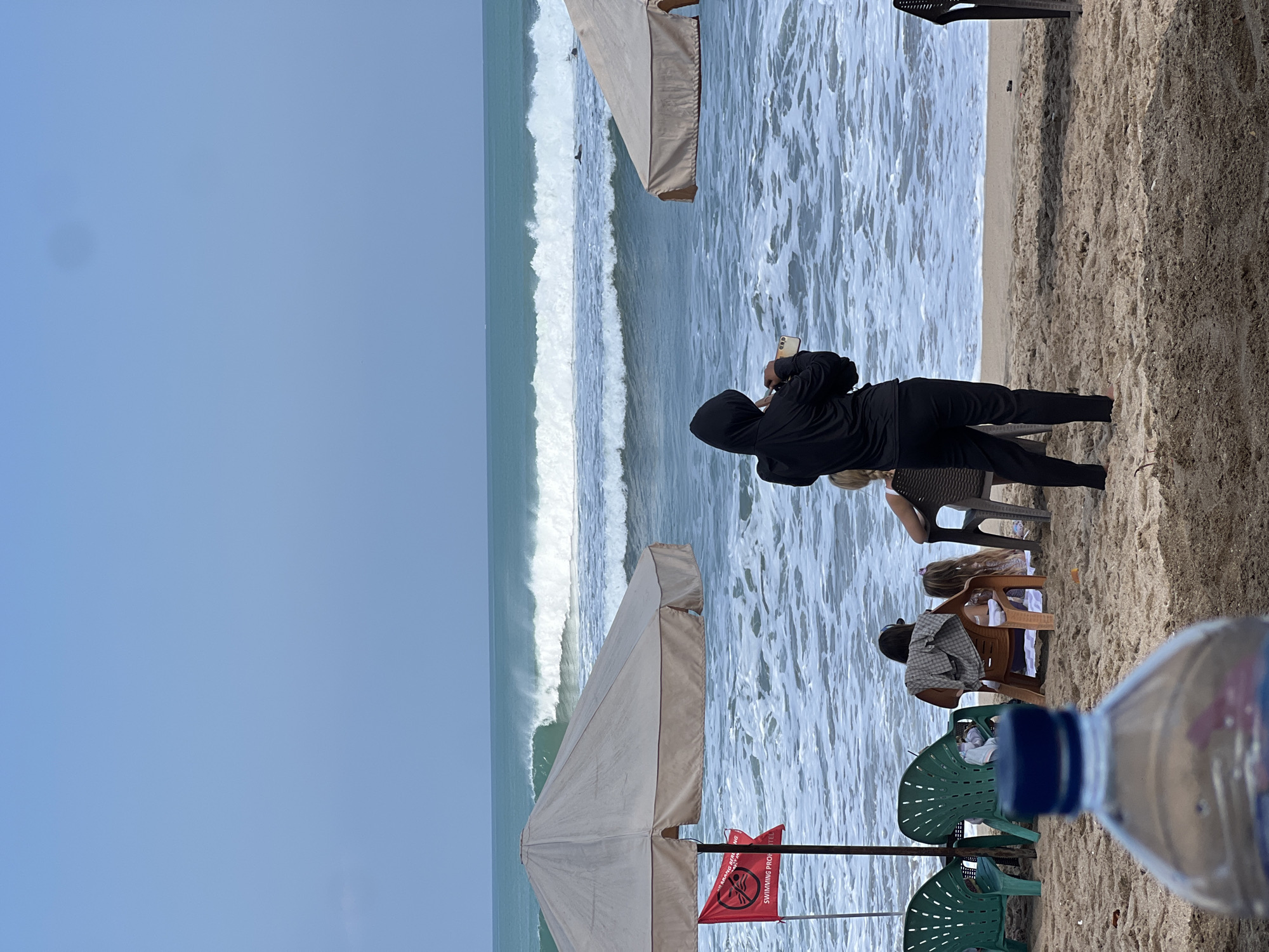 In Kuta, Indonesia, on a sandy beach, a woman in a black hooded jacket stands facing the ocean, holding a phone to take a picture. She is positioned on the sand, with the ocean's waves and white foam in the background. Several beach umbrellas are visible, including a large beige one on the left and another partially visible on the right. A red flag with a prohibition symbol is attached to a pole near the left umbrella, indicating that swimming is not permitted. In the middle ground, several people are seated on plastic chairs on the sand, facing the water. One person is wearing a plaid shirt and has a towel draped over their shoulders. A child is seated next to them. The water is a vibrant turquoise, with visible waves breaking. The sky is clear and light blue. In the foreground, the out-of-focus neck and cap of a blue-lidded plastic water bottle are prominent.