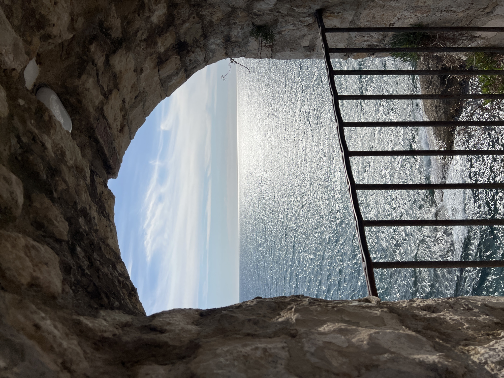 View through a stone archway of the Adriatic Sea and sky, Ulcinj - Ulqin, Montenegro. The left side of the archway contains a light fixture. The right side shows a rusty railing separating a drop to the sea, with small plants and grass growing along the edge. The sea is bright with sunlight. The sky is pale blue with white clouds on the left.