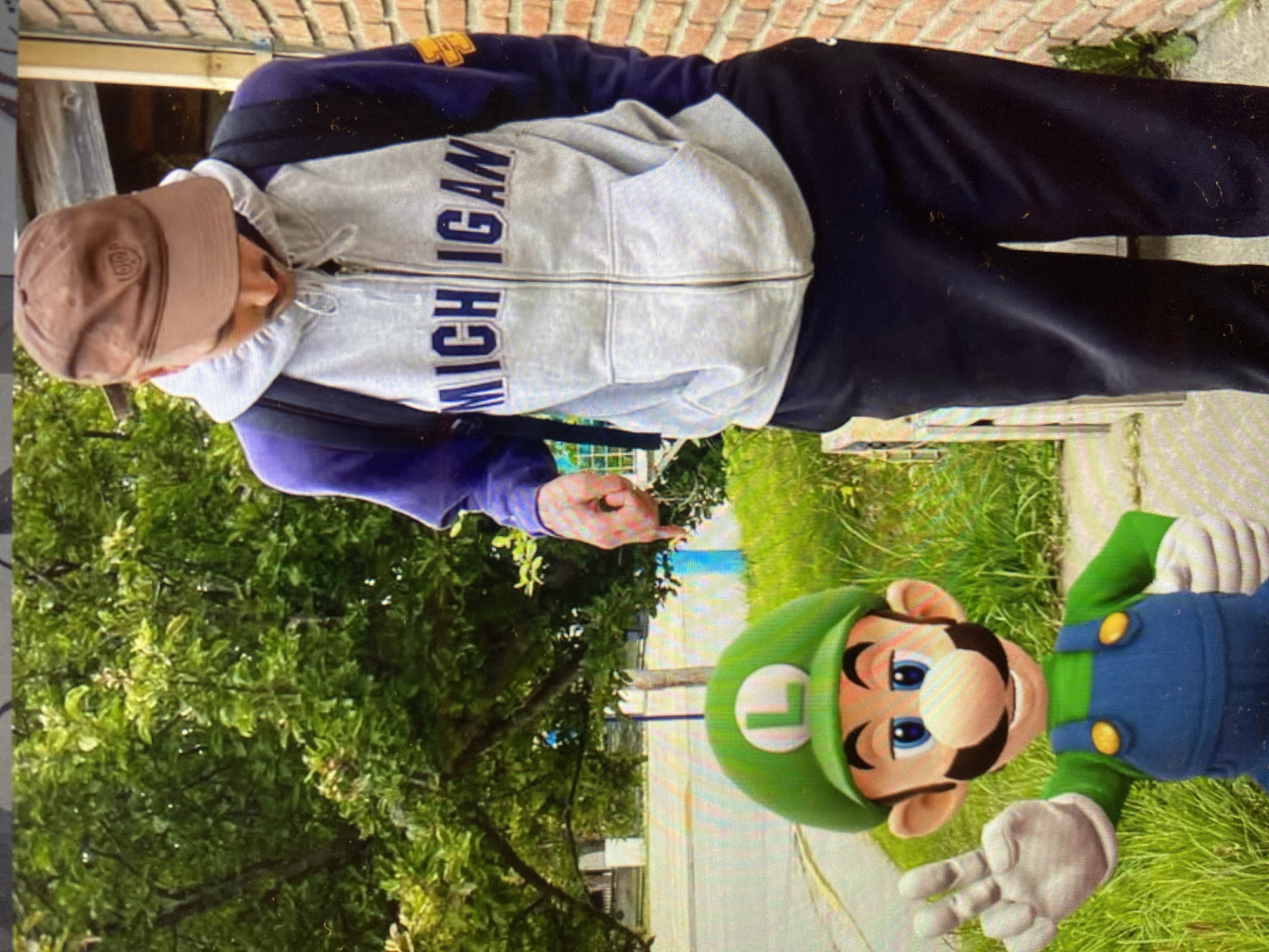 A man in a brown baseball cap, a gray and purple Michigan zip-up hoodie, and black pants stands outside, pointing at a large, cartoonish Luigi character from the Mario franchise. The location is unclear; the background includes a brick wall, green trees, and grass. There is no indication that this photo was taken in the city of Buenos Aires, Argentina.