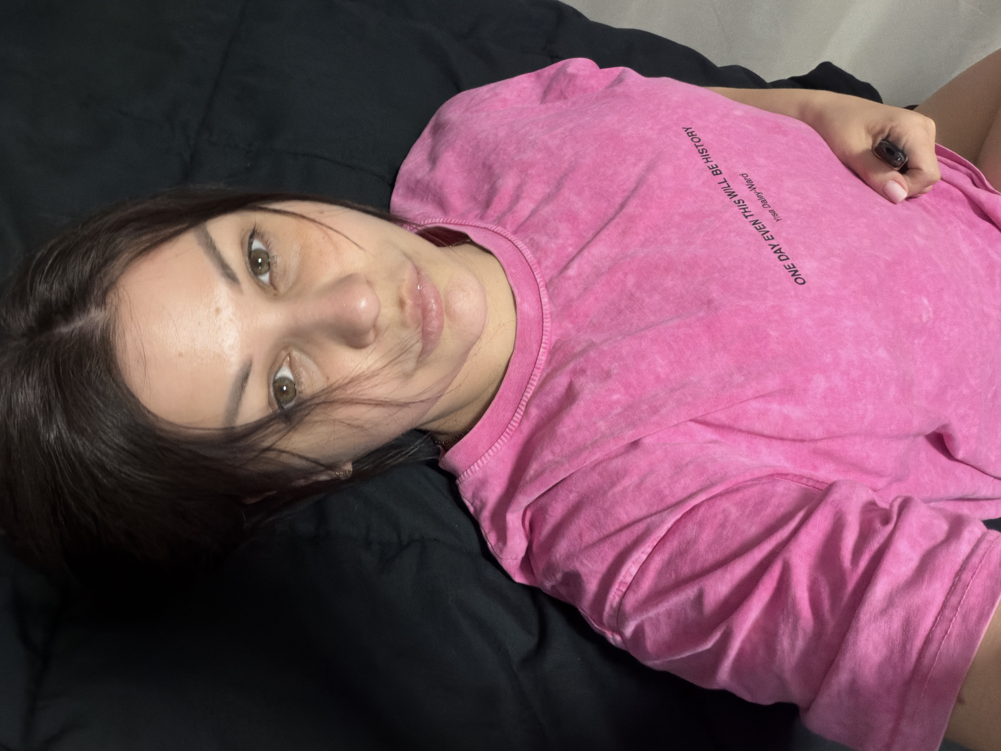 The image depicts a young woman lying on a black surface, possibly a bed or couch. She is wearing a pink t-shirt with text on it. The text is in English and reads "ONE DAY EVEN THIS WILL BE HISTORY Y/rse Daley-Ward". The woman has dark hair and appears to be looking towards the camera. She is holding a dark-colored object in her right hand. The lighting suggests an indoor environment. There is no indication of the location being Sochi, Russia.