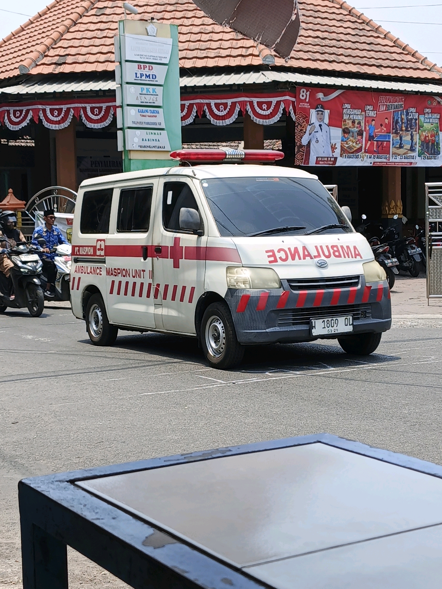 An ambulance with "AMBULANCE MASPION UNIT II" written on its side is parked on a paved road in Ketegan, Indonesia. The ambulance is white with red accents and has a red flashing light bar on its roof. A sign with various logos and text is visible behind the ambulance. In the background, there is a building with a tiled roof and a banner with images and text. Two individuals on motorcycles are also visible on the left side of the image. A dark-colored table is in the foreground.