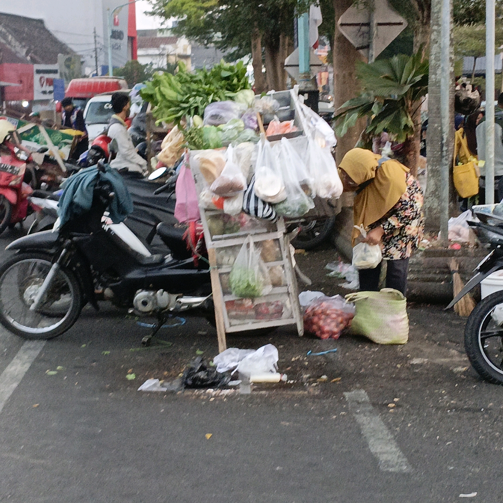 In Salatiga, Indonesia, a street scene unfolds with a woman in a mustard-colored headscarf and a floral-patterned top standing near a mobile market stall. The stall is laden with various produce, including leafy greens and red onions, displayed in plastic bags and baskets. In the foreground, a black motorcycle is parked, its handlebars covered with a green cloth. Various other motorcycles and a red delivery vehicle are visible in the background, suggesting a busy urban environment. The ground is paved with asphalt and appears wet, with scattered debris and fallen leaves. The overall atmosphere suggests a typical day at an Indonesian street market.