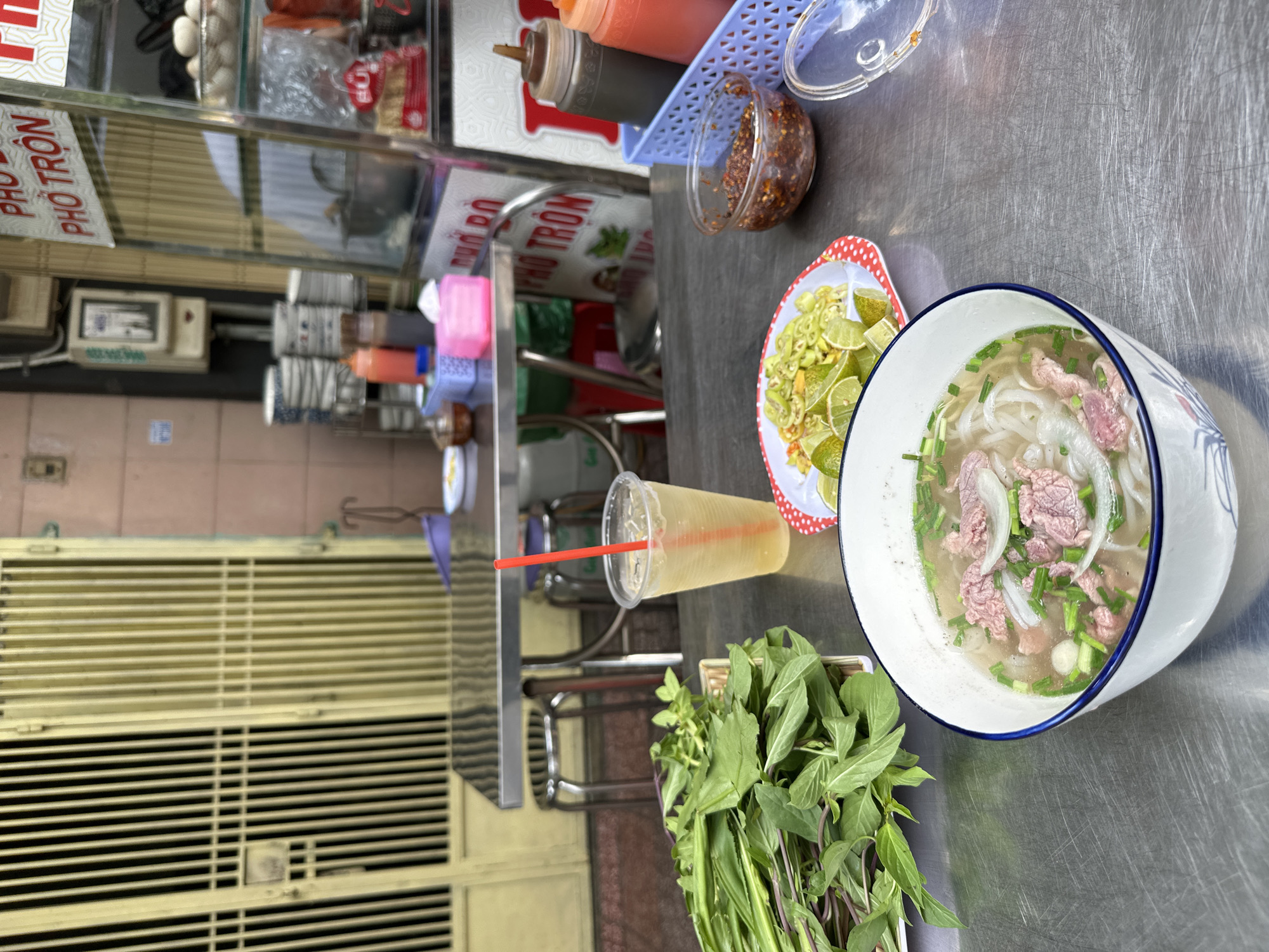 This photo shows a bowl of pho served on a stainless steel table, along with a drink, a dish of limes and chili peppers, and a bunch of basil. The scene is outdoors, with a metal grate and a food cart visible in the background. Based on the food and environment, this photo was taken in Ho Chi Minh City, Vietnam.