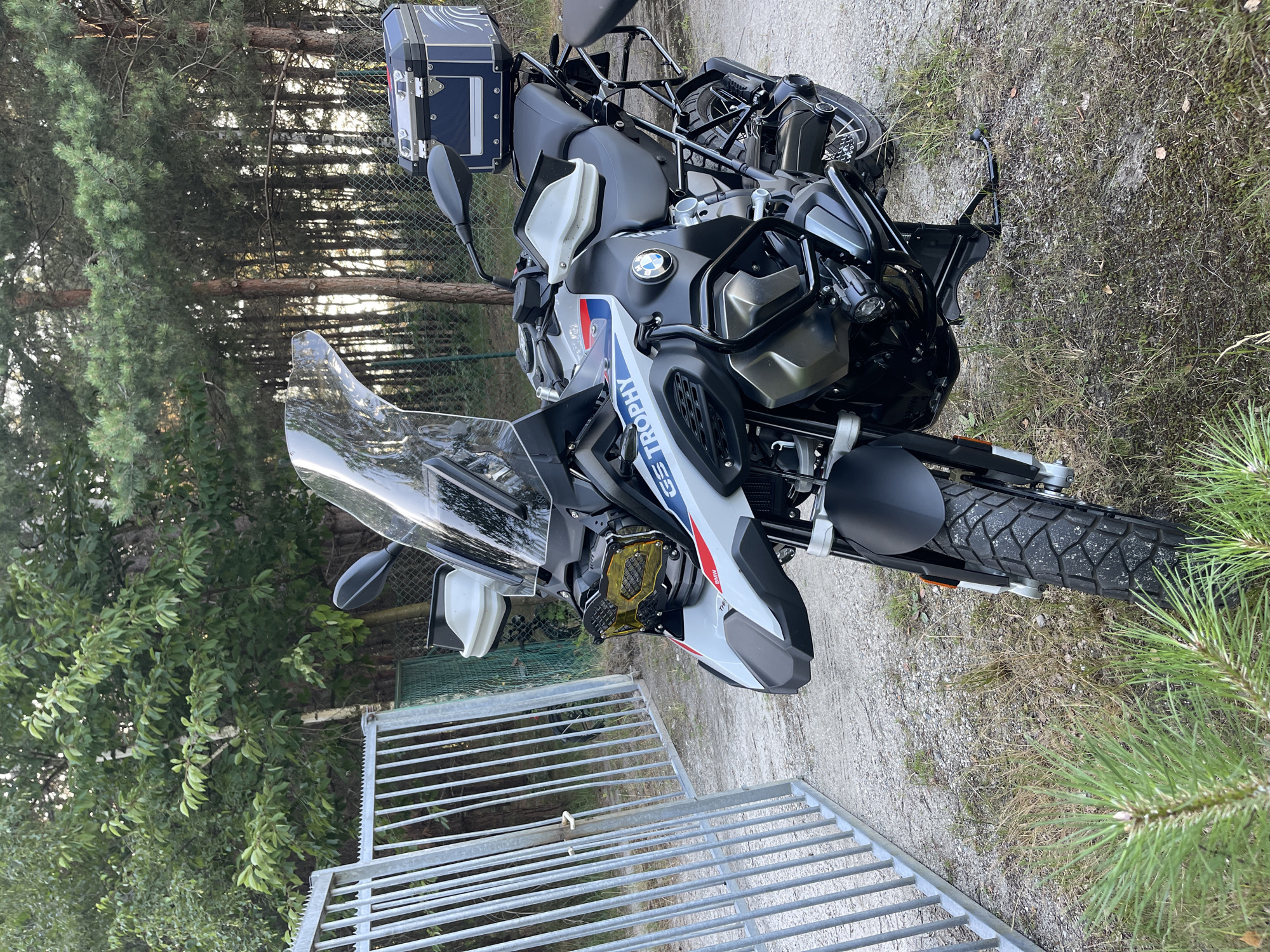 An adventure touring motorcycle, a BMW R 1250 GS Trophy, is parked outdoors on a dirt path next to a metal fence. The motorcycle is predominantly white with blue and red accents, featuring a large windshield, a luggage box on the rear, and protective bars. The environment appears to be a wooded area with trees and dry grass, suggesting a rural or natural setting. The lighting indicates it might be late afternoon or early morning.