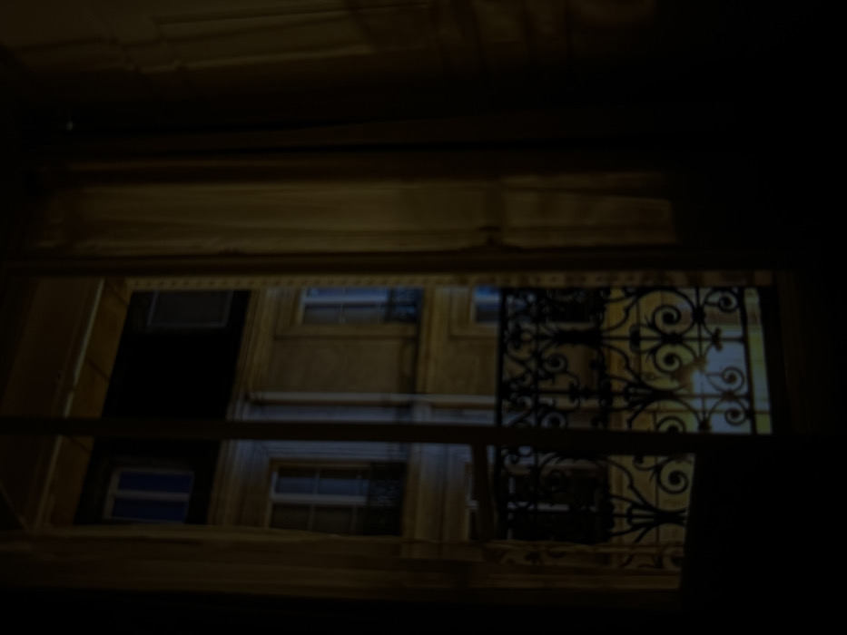 A low-angle shot reveals a building facade framed by a window. The building features symmetrical window placement and ornate wrought iron detail on one section. The image is dark, obscuring details, but hints at a building illuminated by ambient light. The window frame suggests an indoor perspective looking out onto the building across a narrow space.