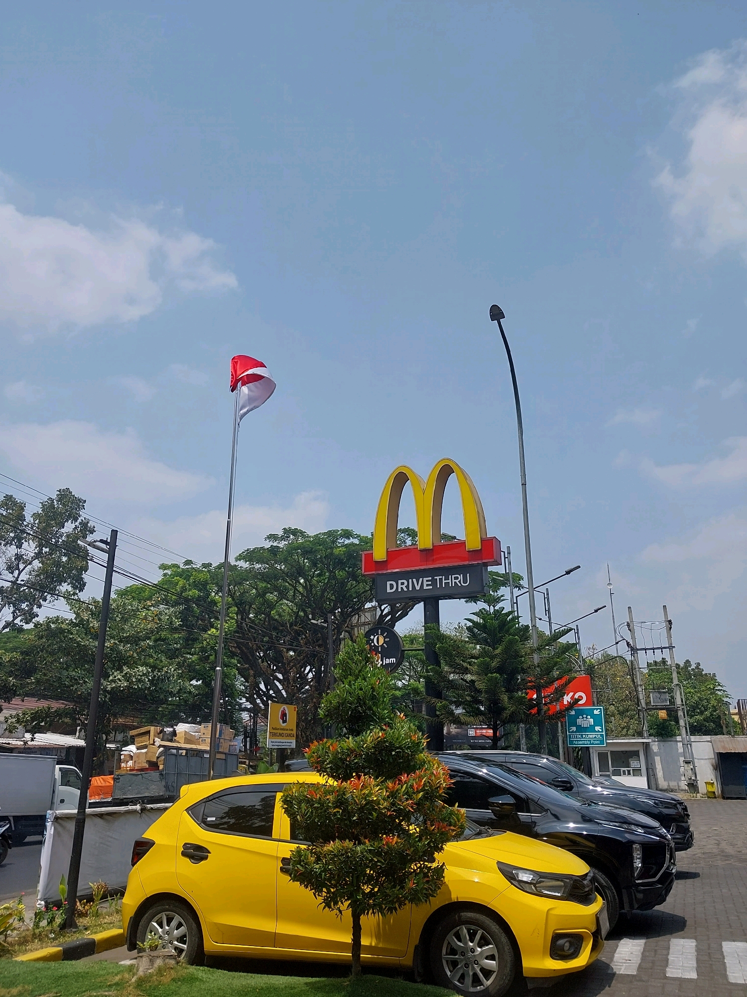 An outdoor, eye-level medium shot captures a bright sunny day with a clear blue sky and scattered clouds, possibly in Malang, Indonesia. The prominent McDonald's "Drive-Thru" sign, featuring the iconic golden arches, stands tall against the sky, with a red base and white lettering. To the left, an Indonesian flag, a horizontal bicolor of red and white, flutters on a flagpole. In the foreground, a vibrant yellow car is parked, its driver's side visible. Behind it, two black SUVs are lined up, suggesting a drive-thru queue. Lush green trees and manicured bushes frame the scene, and utility poles with numerous wires dot the background. A large truck filled with boxes is also visible to the left, hinting at commercial activity. The overall atmosphere is one of everyday urban life on a pleasant day.