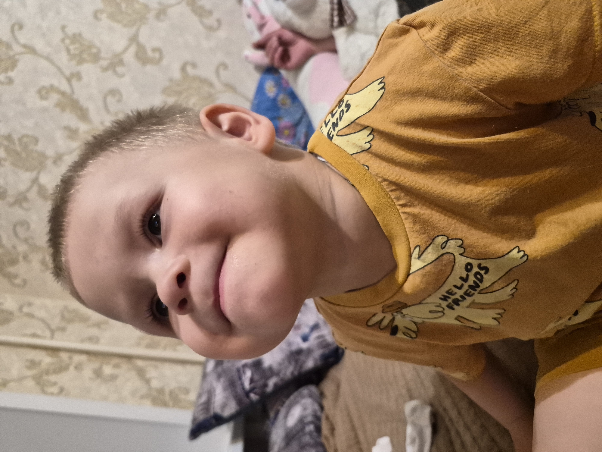 A child, possibly male, with fair skin and short blonde hair is shown from the chest up. The child is wearing a mustard-yellow t-shirt with a graphic design of a cartoon dog and the words "HELLO FRIENDS" printed on it. The child's head is tilted upwards and to the left, with cheeks puffed out and a slight smile. The background consists of a textured wallpaper with a floral pattern in beige and cream tones. To the left of the child, a dark-colored pillow with a grey pattern is visible, resting on a brown surface, possibly a couch or bed. Further in the background, to the right, a brightly colored pillow with pink and blue elements is partially visible, along with what appears to be a stuffed toy. The lighting suggests an indoor setting, possibly a home environment in Chaikovsky, Russia.