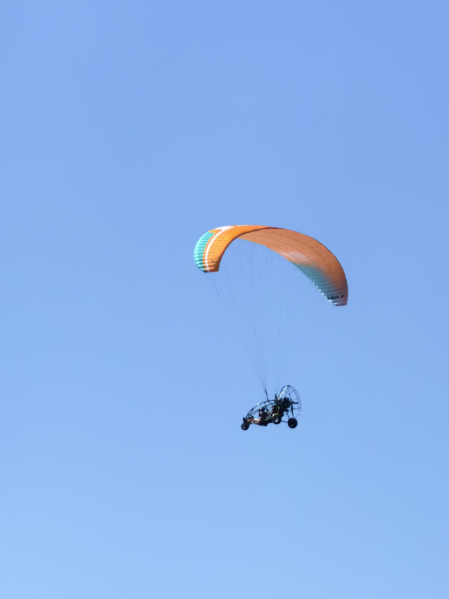 A powered paraglider is in flight against a clear blue sky. The paraglider has an orange and light blue wing and a wheeled frame carrying two people. The paraglider is positioned in the lower right quadrant of the frame. The image is taken from below, looking upwards. The specific location in Shikhan, Russia cannot be determined from the image.