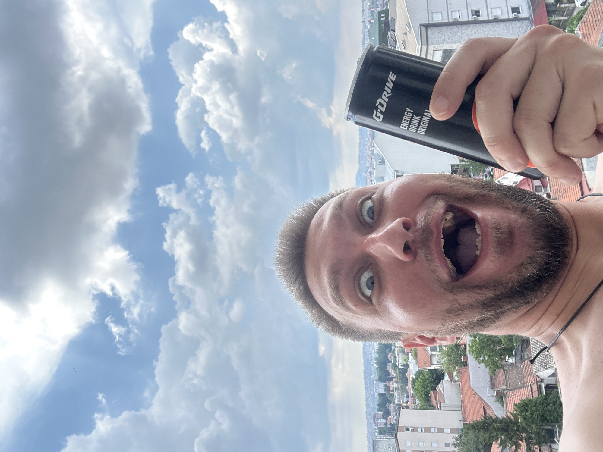 The image is a selfie taken from a low angle, showing a man with wide, surprised eyes and an open mouth, holding a black can of "G-DRIVE ENERGY DRINK ORIGINAL" in his right hand. He is shirtless and appears to be on a balcony or rooftop, as evidenced by the railing visible at the bottom of the frame and the urban cityscape in the background. The cityscape features several buildings with red-tiled roofs and various architectural styles, suggesting a residential area within the Zvezdara Urban Municipality of Serbia. The sky above is filled with dynamic, white and grey clouds against a bright blue background, indicating it is daytime. The man's expression and the selfie-style composition suggest a spontaneous or playful moment.