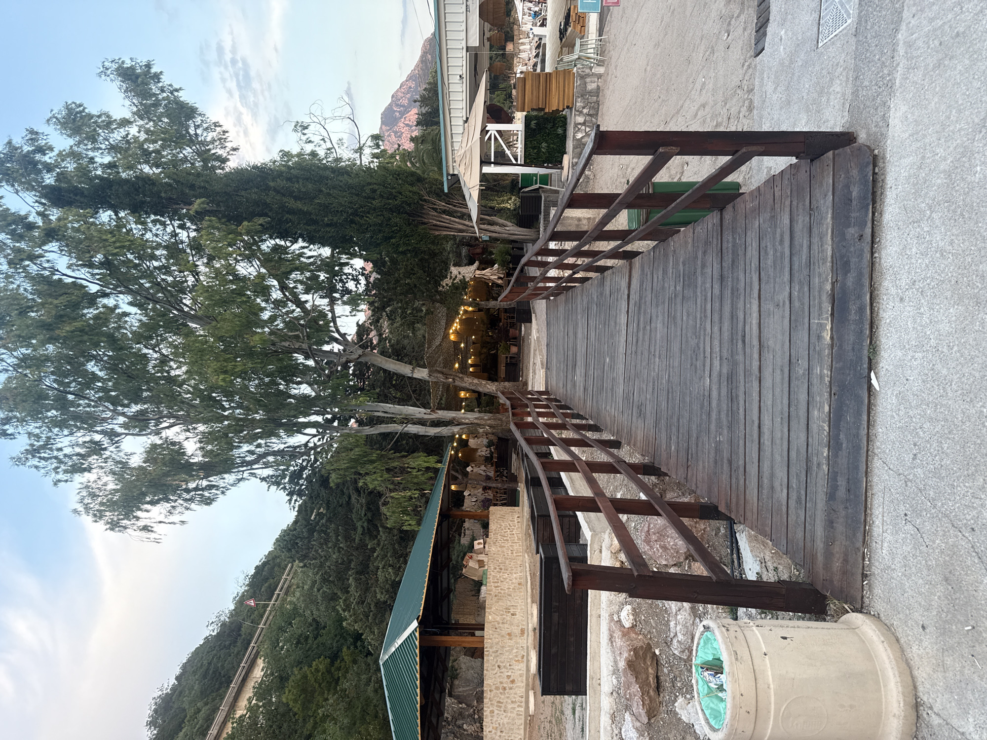 The image depicts an outdoor scene at dusk, likely in a Mediterranean coastal setting, given the location context of Kamenovo, Montenegro. A wooden bridge with dark brown railings curves into the frame from the right, leading towards a softly lit outdoor dining area. The bridge is constructed from weathered wooden planks.
On the left side of the bridge, a large tree with a thick trunk and abundant green foliage dominates the view, its branches reaching towards a pale sky with wispy clouds. Beneath the tree and to the left of the bridge, a covered outdoor seating area is visible, illuminated by strings of warm, yellow lights. Tables and chairs are arranged within this area, suggesting a restaurant or cafe.
In the background, behind the outdoor seating, a building with a light-colored facade and a green-tiled roof is partially visible. Beyond this, a rocky, tree-covered hillside rises, with a hint of a mountain peak in the distance. The ground appears to be a mix of sand and stone. A beige trash receptacle with a green liner is situated near the edge of the concrete pathway on the right side of the image. The overall atmosphere is serene and inviting, with the warm lighting creating a cozy ambiance as daylight fades. No visible text is present in the image.