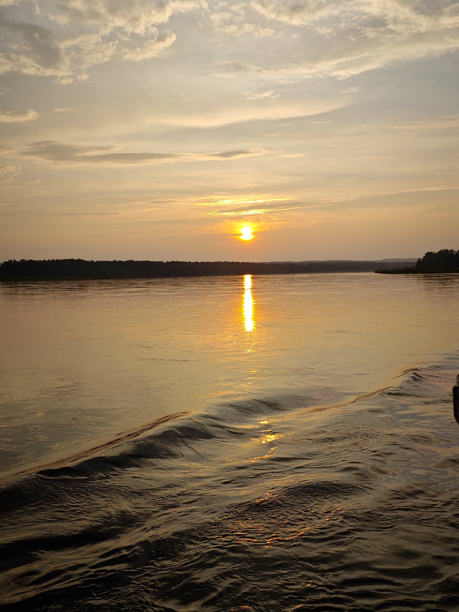 A serene sunset over a wide body of water, likely a river, is depicted in this horizontal landscape photograph. The sun, a bright orb of golden light, is positioned slightly left of the center of the frame, casting a shimmering reflection across the water's surface. The sky is filled with wispy clouds tinged with hues of orange, yellow, and pale blue, suggesting the late afternoon or early evening. A dark, tree-lined silhouette of the shore stretches across the horizon, interrupted by a distant, low-lying landmass on the right. The foreground of the image shows ripples and waves on the water, indicating movement, possibly from a boat. The overall scene conveys a sense of tranquility and natural beauty. The location is described as Maloe Nakhval'skoe, Russia, though this cannot be confirmed from the image alone. There are no people or specific objects identifiable in the scene beyond the natural elements.