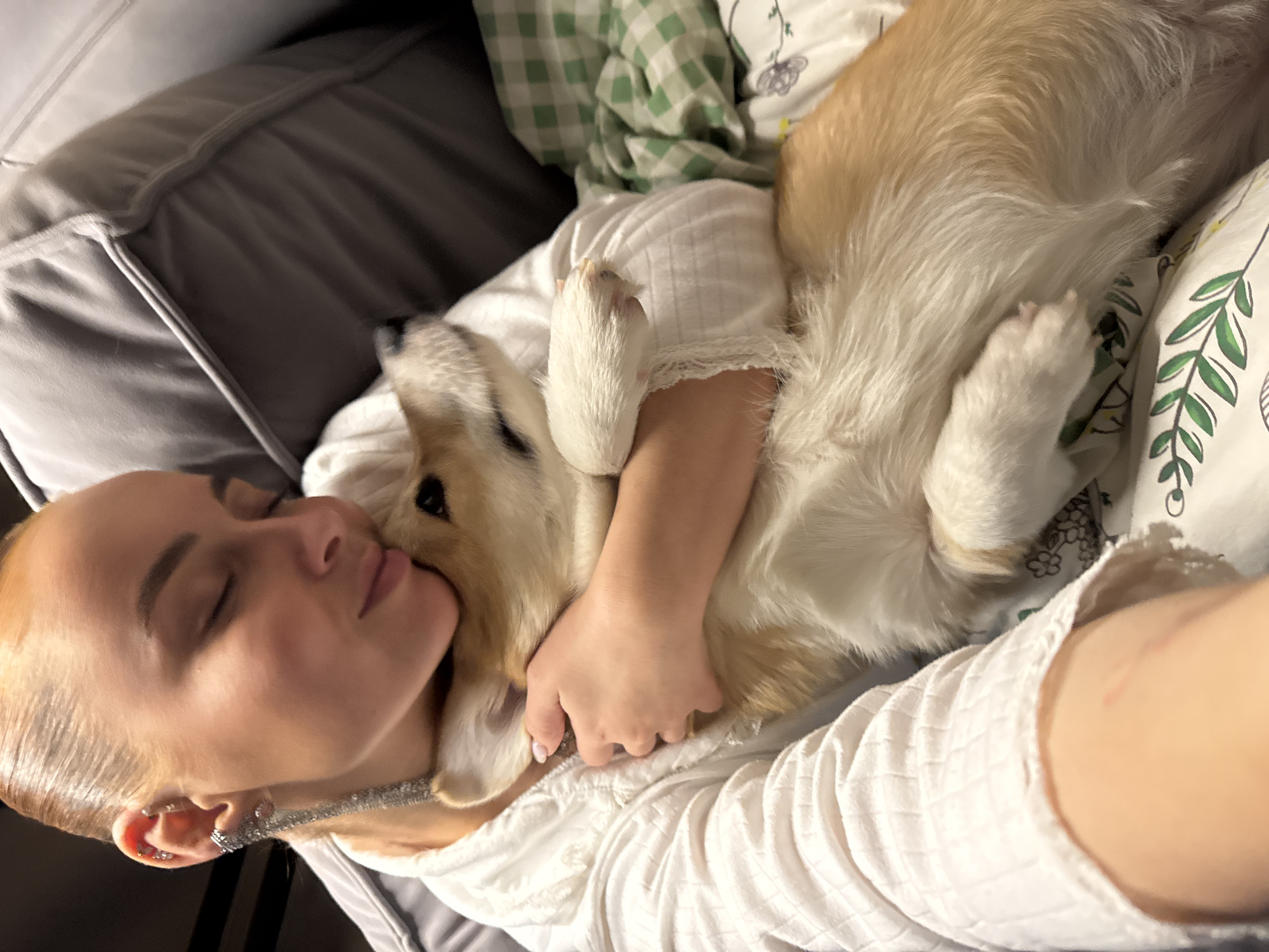 An overhead, slightly Dutch-angled shot captures a woman and a Corgi dog cuddling intimately indoors. The scene depicts a moment of peaceful rest or sleep, likely on a bed or a comfortable sofa.
The woman, positioned on the left side of the frame, has light blond hair pulled back, showcasing her face in profile with her eyes gently closed. She wears a white, ribbed long-sleeved top. Notable details include multiple piercings on her left ear, including an elaborate ear cuff and a shimmering, chain-like earring that drapes towards her neck. Her left arm is seen tenderly embracing the dog.
The Corgi, with its distinctive light brown and white fur, is nestled against the woman's face and upper body. Its head is tucked comfortably near her chin, and one of its paws rests on her arm, suggesting a deep bond and comfort. The dog also appears to be either sleeping or in a state of deep relaxation.
The setting includes soft furnishings. To the left, a large grey upholstered cushion or headboard is visible. To the right and behind the subjects, there are soft textiles including a green and white checkered blanket or sheet, and a white pillow adorned with a green leaf pattern.
The image conveys a strong sense of affection, warmth, and tranquility. The soft, ambient lighting suggests an indoor setting, possibly during the daytime or with artificial light. There is no visible text within the image. The location is Chaikovsky, Russia.
