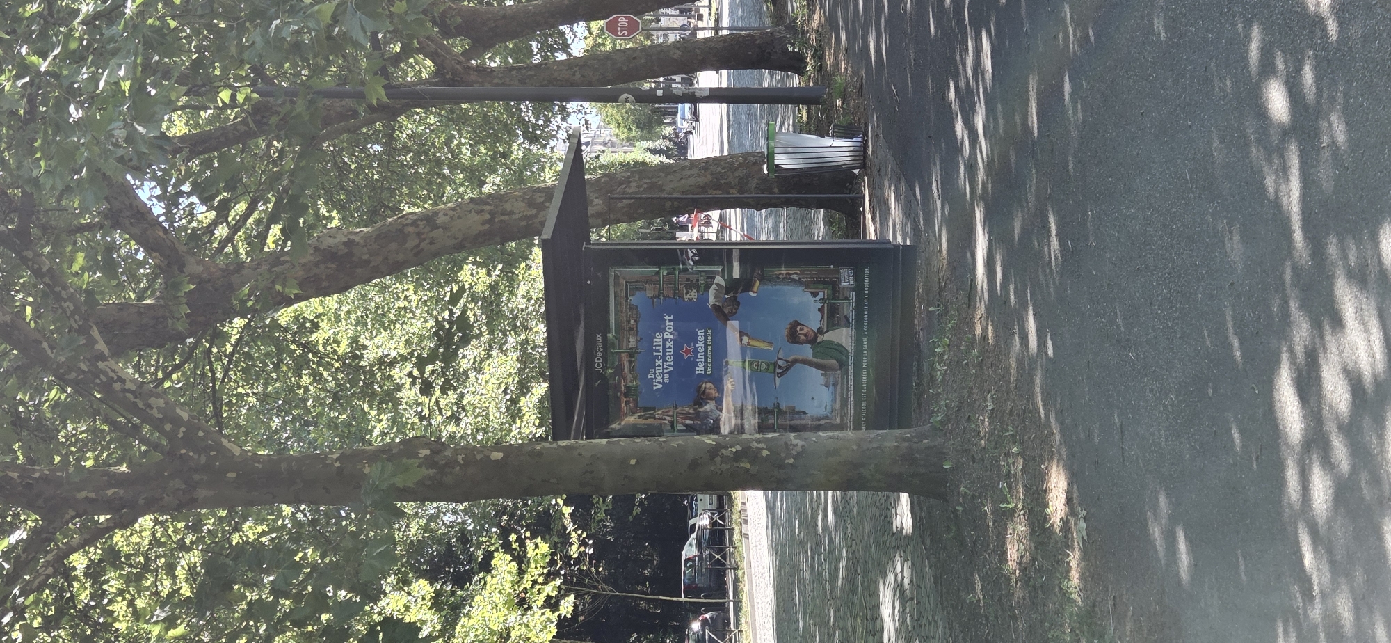 The image shows a bus stop advertisement featuring a Heineken beer ad. The ad has text that reads "JCDecaux" at the top, and "Du Vieux-Lille au Vieux-Port" below that. The Heineken logo is also visible, along with the slogan "Une même étoile". The ad shows a man holding a tray with a Heineken bottle and glass. The bus stop is located on a street with trees lining the sidewalk. In the background, there are cars and buildings. A stop sign is visible on the right side of the image. The scene is set in Strasbourg, France, as evidenced by the text on the advertisement that refers to locations within the city. The lighting suggests it is a sunny day, casting shadows from the trees onto the ground.