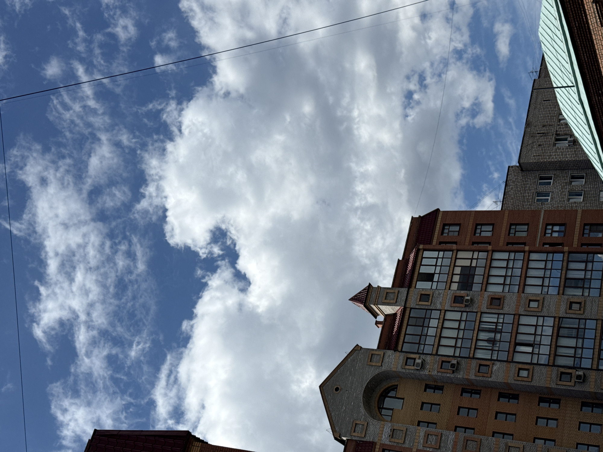 The image shows a building in Krasnoyarsk, Russia, viewed from below against a cloudy sky. The building's facade is brick with numerous windows. A power line is visible in the upper left. The building appears to be multi-storied with architectural details, including gables and decorative brickwork. The sky is blue with scattered white clouds.