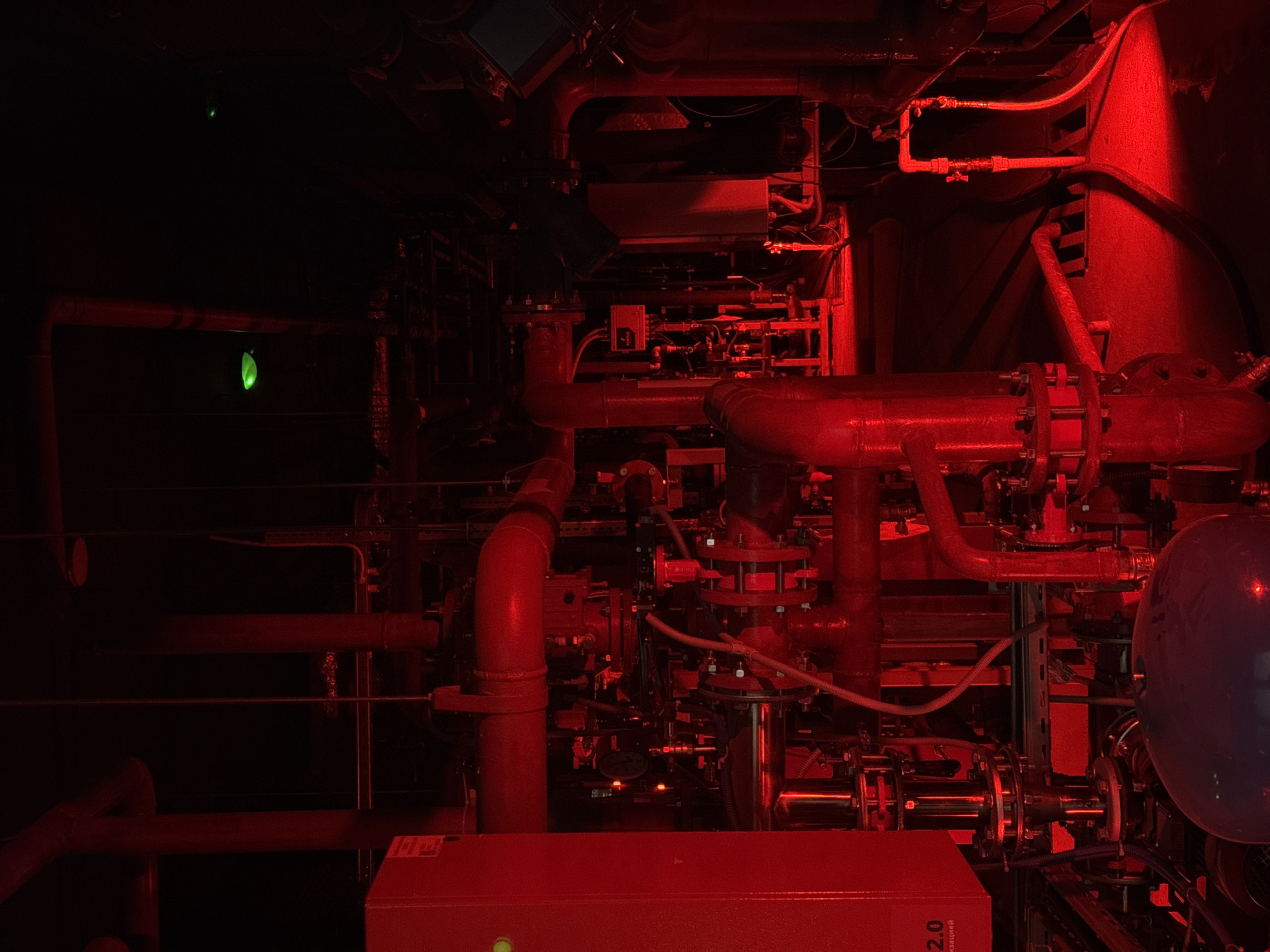 The image presents a dense network of pipes, valves, and industrial equipment, illuminated by a predominant red light, creating a stark and dramatic atmosphere. The equipment appears to be situated within a confined space, possibly an engine room or utility area. A blue-tinted sphere is visible on the right. Near the bottom left, a red box with a green indicator light and the partially visible inscription "2.0" is present. In the top left, two glowing green objects are visible. Given the specific reference to Tyumen, Russia, the image likely depicts a component of the city's infrastructure or an industrial facility. The image shows no people, no movement, no actions, no obvious interactions. The environment is dark and industrial.