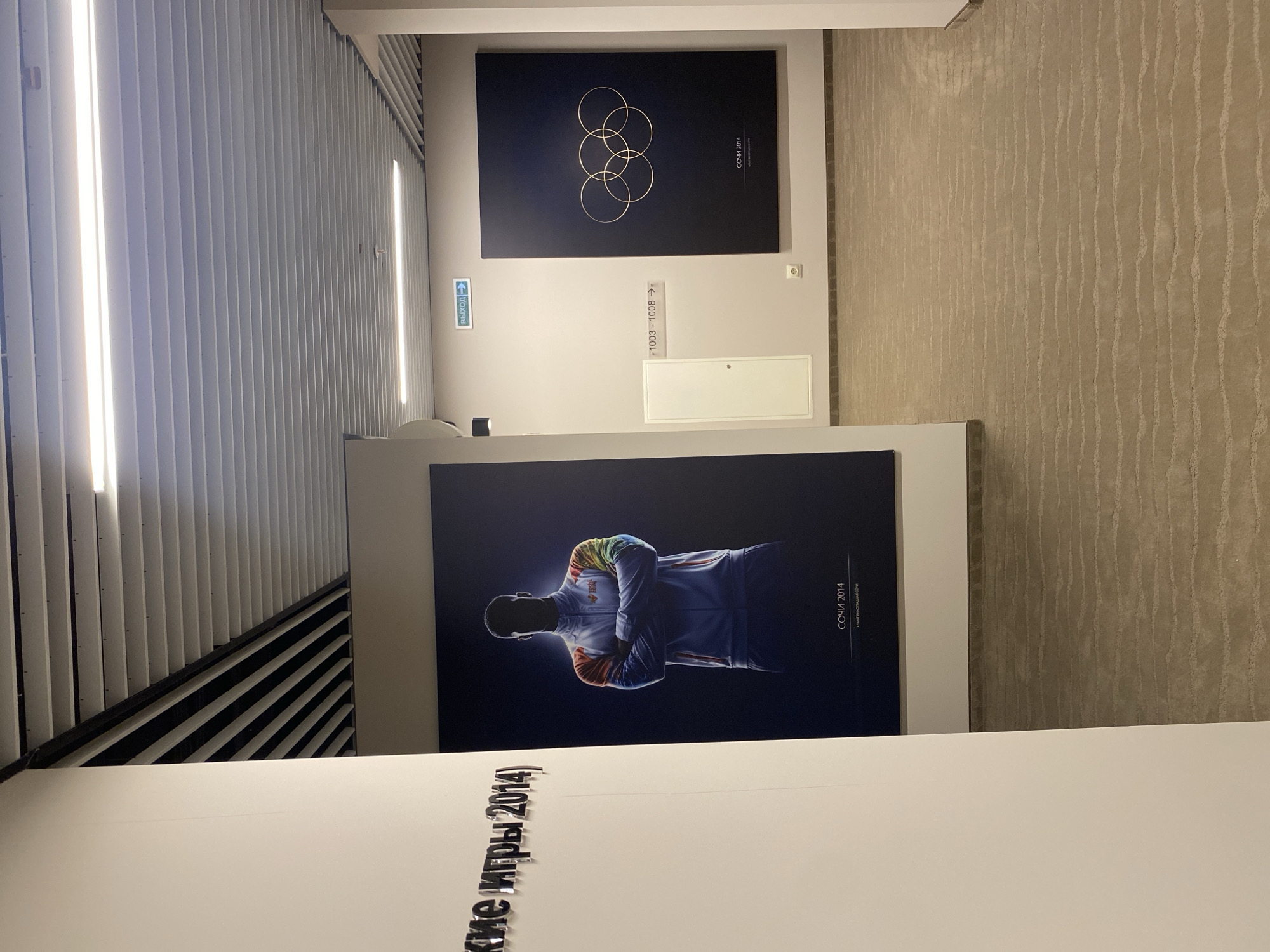 This image shows a hallway in Sochi, Russia, featuring displays related to the 2014 Winter Olympics.
The hallway has vertically aligned white slats on the left. An exit sign in Russian is visible on the wall. Two framed artworks are displayed. The first artwork contains a black background featuring the Olympic rings with the words "SOCHI 2014" in smaller text. The other artwork is a dark photograph of a man in athletic gear with the Sochi 2014 emblem, standing with crossed arms. Below the photograph, the words "Зимние игры 2014" are visible, spelling out the Russian words for "Winter Games 2014".
The floor is covered in patterned beige carpet. The wall adjacent to the hallway is painted light gray and features room number signage and a small white storage panel.