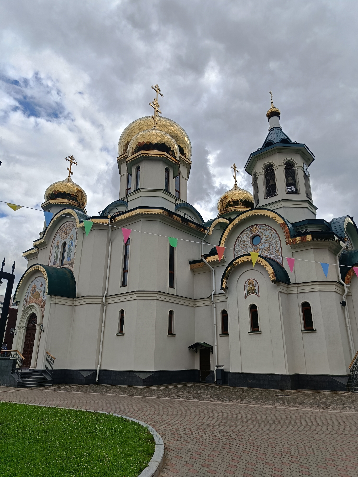 The image shows an exterior view of the Holy Trinity Cathedral of Alexander Nevsky Lavra in Saint Petersburg, Russia. The cathedral is a large, white building with multiple golden domes topped with crosses. The sky is overcast with clouds. Prayer flags in various colors are strung across the front of the building. The foreground features a paved walkway and a green lawn.