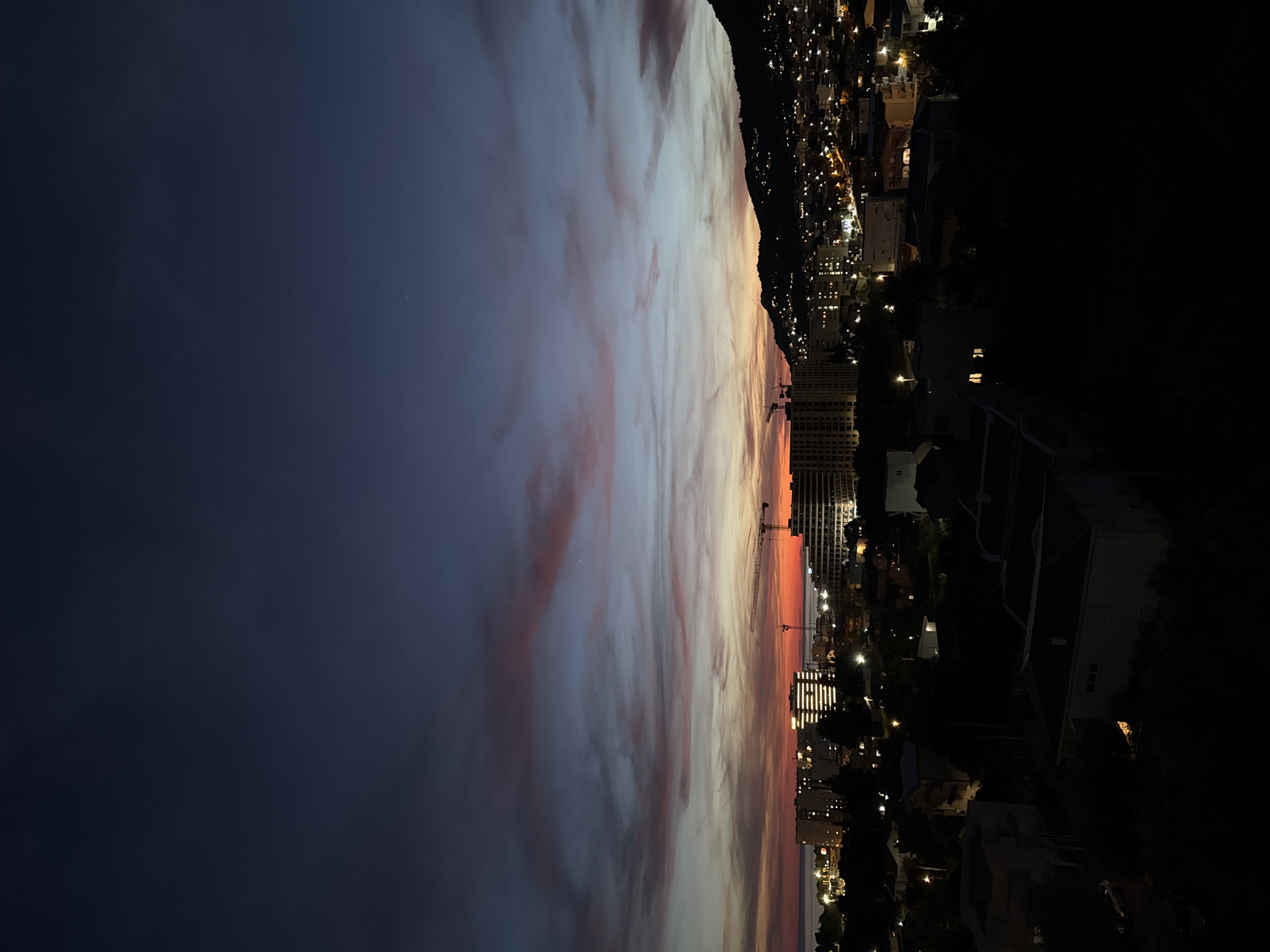 This is a nighttime, high-angle, full shot of a coastal city, likely in the vicinity of Šušanj, Montenegro, as the sun has set and the sky is filled with remnants of twilight colors and clouds. The cityscape is illuminated by numerous artificial lights from buildings and street lamps, creating a warm glow against the darkening sky. Several multi-story buildings are visible, some appearing to be under construction, indicated by the presence of a prominent construction crane in the middle ground. The sky transitions from a deep navy blue at the top to softer hues of pink and orange closer to the horizon, where a faint line of the sea or a large body of water is suggested. Hills or mountains form a dark silhouette against the sky on the right side of the image, dotted with more lights from houses or structures. The foreground consists of darker silhouetted structures, possibly residential buildings or dense foliage, contributing to the overall nighttime atmosphere. There are no people or animals clearly visible in the frame. The scene depicts a developing urban environment at dusk.