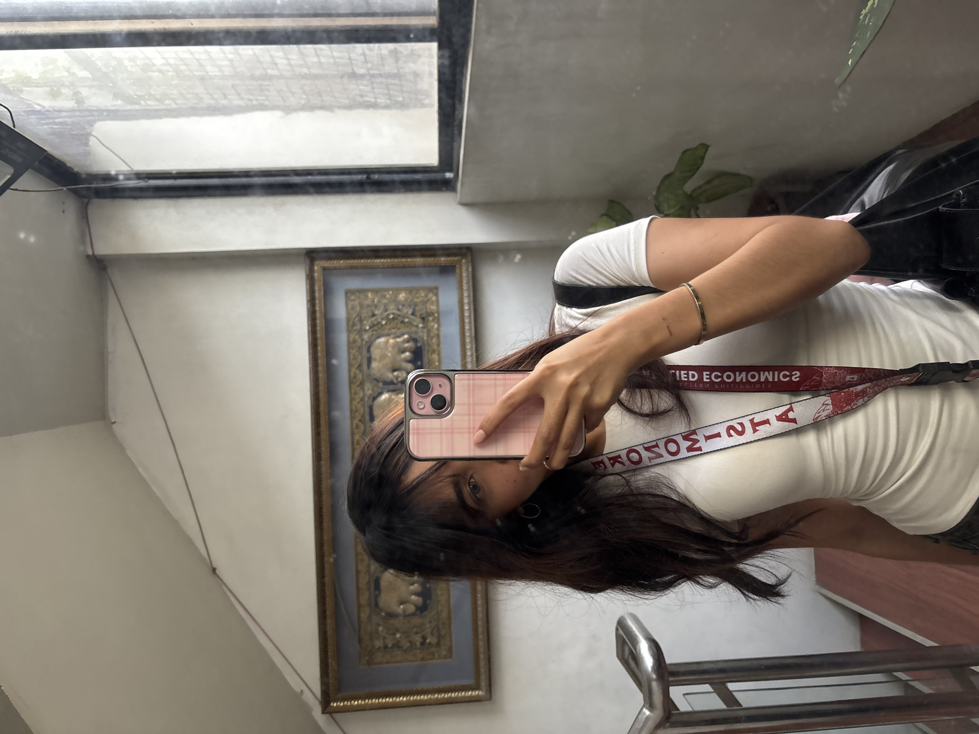 A person is taking a selfie in a mirror. The person is holding a pink-cased smartphone in their right hand. The person is wearing a white T-shirt, a bracelet on their right wrist, and a lanyard around their neck. The lanyard is red and white, with the words "APPLIED ECONOMICS" printed on it in reverse. Behind the person is a framed artwork of an elephant with a golden background. There is a window above the artwork. A plant is visible in the upper right corner of the image. There is a metal railing on the bottom-right corner of the photo. The reflection in the mirror indicates that the photo was taken indoors, possibly in a building in Davao City, Philippines.