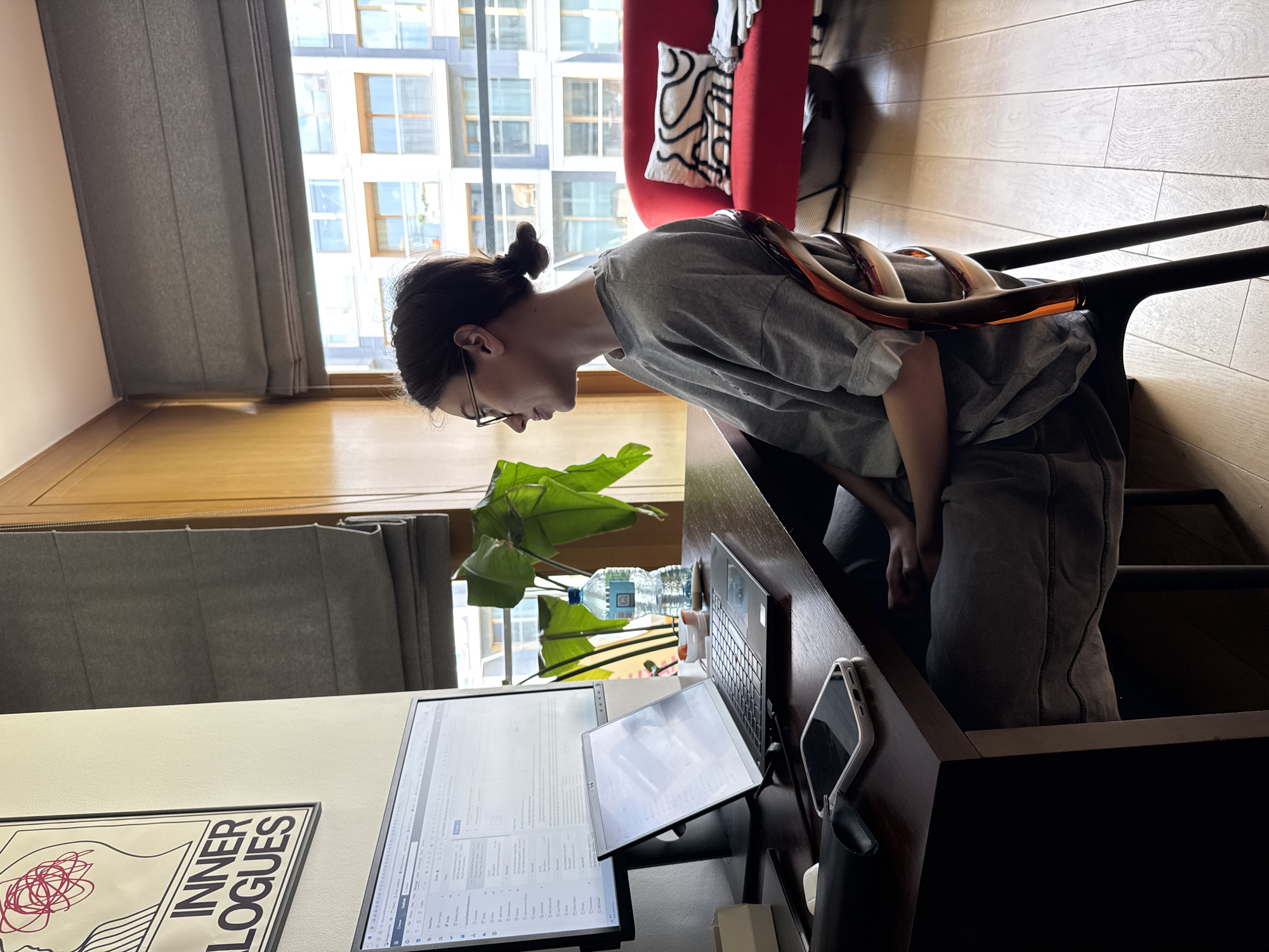 A person sits at a desk in a room with a large window, likely an apartment. The person is facing a computer setup with two monitors. A laptop and a mobile phone are also on the desk. A houseplant and two bottles of water are visible behind the monitors. The person is wearing a gray t-shirt and gray pants. A red chair is visible in the background. A framed poster with the words "INNER LOGUES" is on the wall near the computer setup. The window reveals a cityscape with multiple buildings, suggesting the location is an urban setting.
