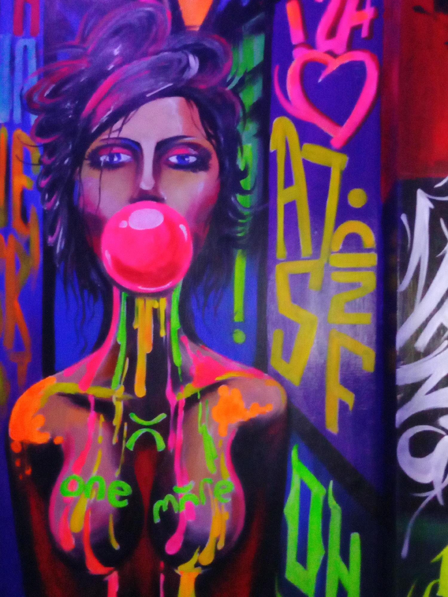 The image is a vibrant, colorful painting depicting a woman blowing a bubblegum bubble. The woman has dark hair with magenta highlights, fair skin, and blue eyes. A large, pink bubble obscures part of her face. Colorful paint appears to be dripping down her neck and chest.
The words "one more" are painted on her chest in green. Graffiti is present in the background, with the letters "A7" and a heart prominently displayed. The word "ON" is also visible.
Based on the style and subject matter, it is likely a piece of street art or graffiti art, possibly located in Nha Trang, Vietnam.