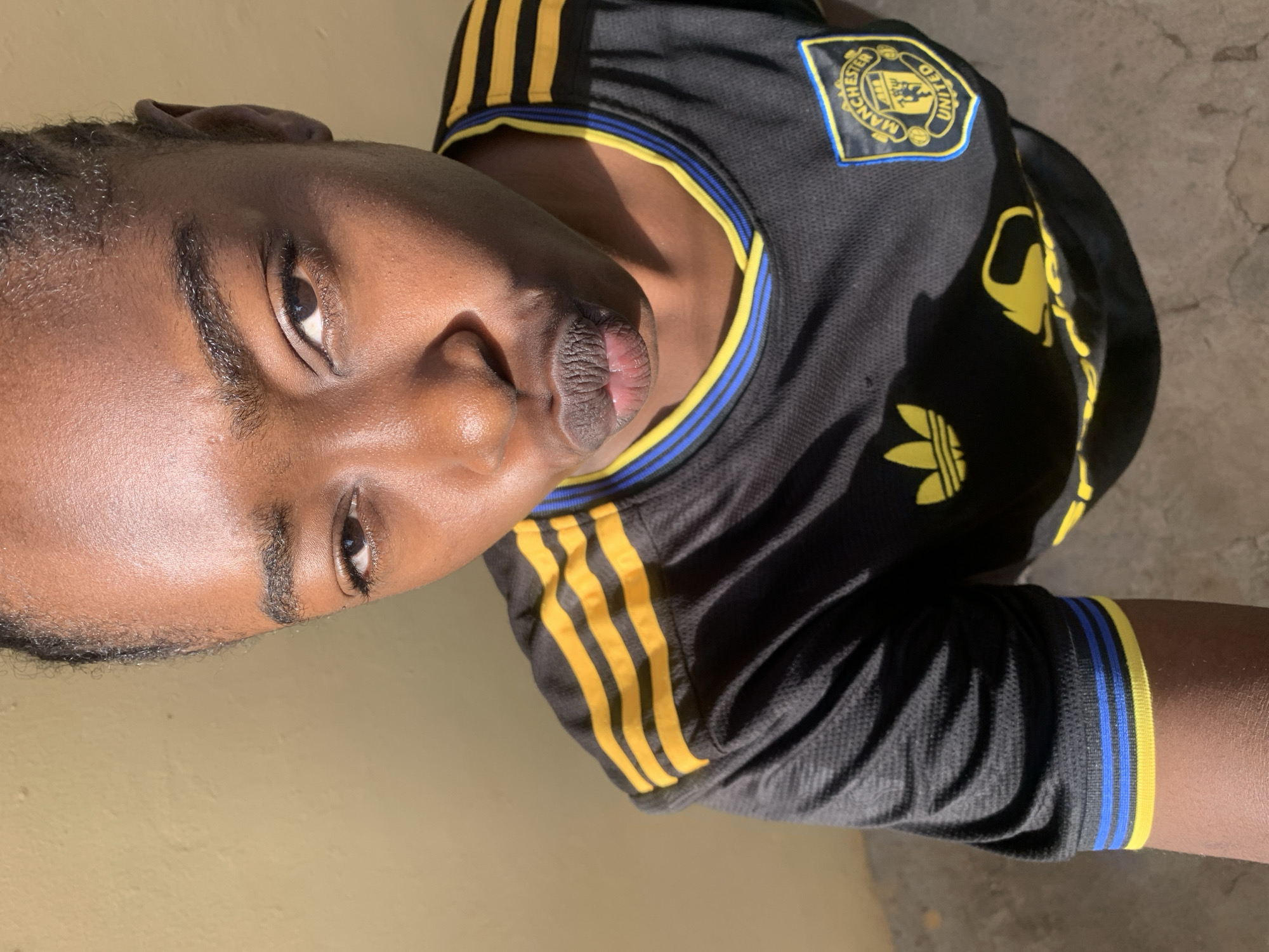 A young Black woman with braided hair is shown from the chest up, looking directly at the camera with her lips pursed in a kissing gesture. She is wearing a black Manchester United football jersey with yellow and blue accents. The jersey has the Adidas logo on the left chest and the Manchester United crest on the right. The background is a plain, textured yellow wall. The lighting suggests it is daytime, with sunlight casting shadows on the wall and the woman's face and jersey. The image appears to have been taken in an outdoor or semi-outdoor setting, possibly a street or a courtyard, in Rafin Par, Nigeria.