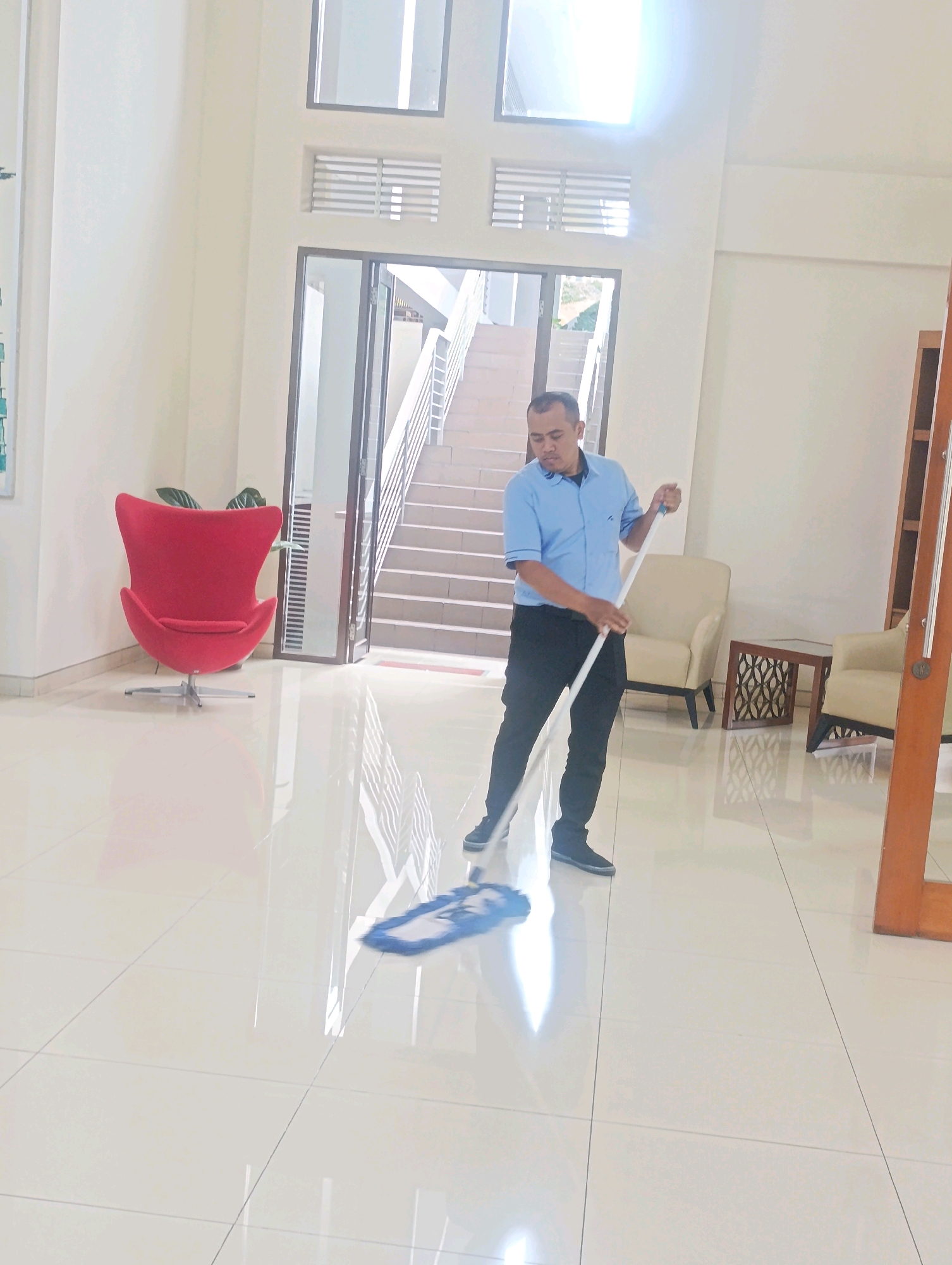 A man in a blue short-sleeved collared shirt and black pants is mopping a very clean, shiny white tiled floor in what appears to be a modern building lobby. He is holding a white mop with a blue mop head, which is actively being used on the floor. The man is positioned in the center of the frame, facing towards the right. In the background, there is a staircase leading up with a railing. To the left, a bright red egg chair is visible, and to the right, there are two cream-colored armchairs and a wooden table with an intricate pattern. Large windows are present on the back wall, allowing natural light to enter, creating reflections on the polished floor. The overall environment suggests an interior space within a public building or a commercial establishment. There is no specific indication of the city of Cihanjuang, Indonesia, within the image itself.