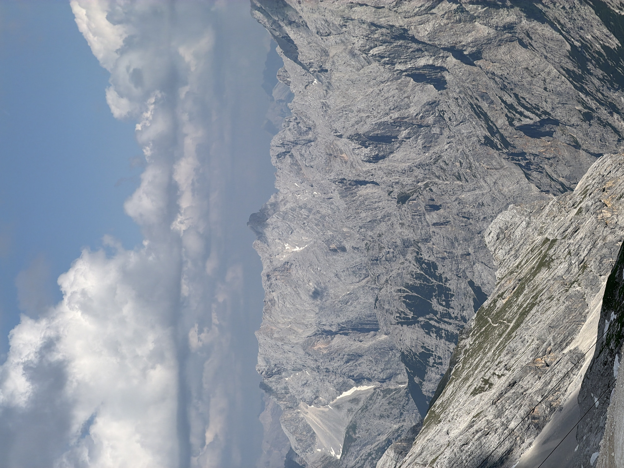The image depicts a panoramic view of a rugged mountain range under a partly cloudy sky. The mountains are predominantly rocky with steep slopes and jagged peaks, displaying shades of gray and white due to rock formations and patches of snow. Some areas show sparse vegetation, appearing as green streaks on the slopes. In the foreground, a steep, rocky incline is visible, with what appear to be cables or tracks running diagonally down its side, suggesting a cable car or ski lift system. The sky is a vibrant blue with scattered white and gray clouds. The overall scene is one of a vast, natural alpine landscape. This scene is consistent with the Zugspitze area near Garmisch-Partenkirchen, Germany, a popular destination for its mountainous terrain and recreational activities. There are no people or animals visible in the image.