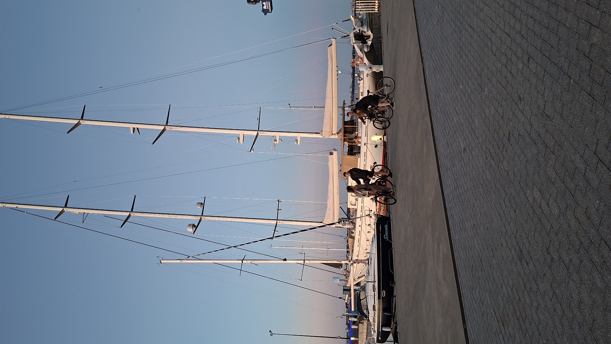 The image depicts a waterfront scene in Tallinn, Estonia, likely during the evening given the soft lighting. Prominently featured are several large sailboats docked at a pier. In the foreground, a paved walkway leads towards the boats. A group of four individuals is present, each on a bicycle, some engaged in conversation or pausing their ride. One smaller dark blue sailboat, named "Scorpio," is docked to the left, while larger white sailboats are positioned to its right, with their masts reaching high into the clear blue sky. The environment suggests a public area, possibly a marina or promenade, with the city skyline visible in the distance.