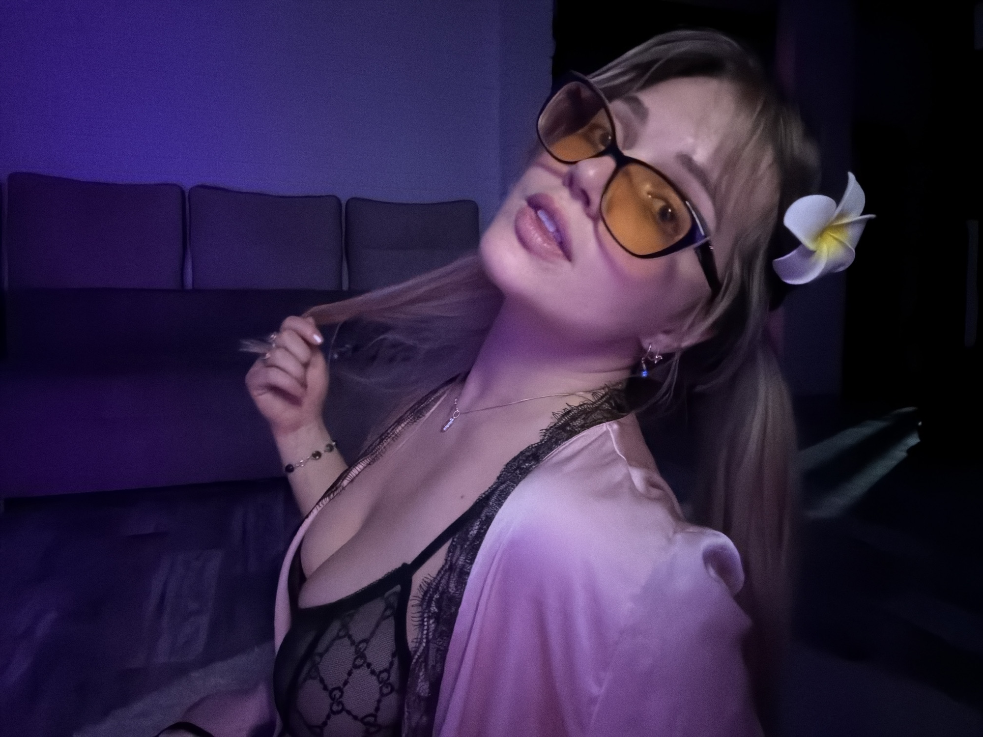 A young woman with blonde hair, wearing glasses, a pink robe, and a lace top, poses in what appears to be an indoor setting. She has a flower in her hair. The background includes a couch and some dark areas. This image does not provide definitive information about the location being in the city of Большая Шильна, Russia.