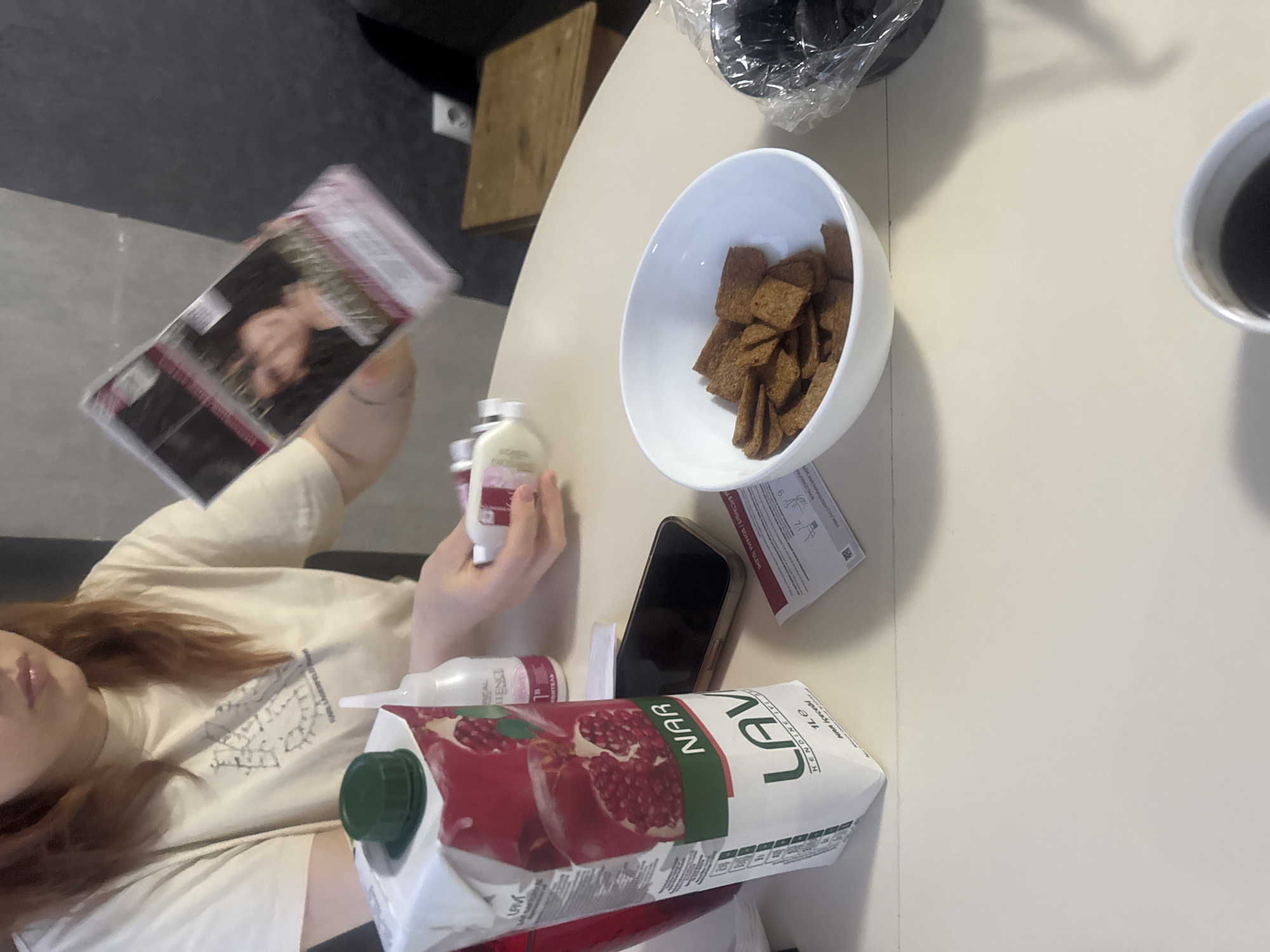 A medium overhead shot features a portion of a woman holding objects on a light-colored table, possibly indoors.
The woman, partially visible on the left side of the frame, has red hair and wears a light-colored t-shirt. She holds a hair dye box in her left hand, and in her right hand, she holds a small white bottle.
Several items are arranged on the table. To the right of the woman, there is a package of pomegranate juice, and a white bottle of hair dye, a black smartphone, a white bowl containing snack crackers, a cup of coffee, and a leaflet.
The location is potentially indoors in a private residence but city location cannot be verified.