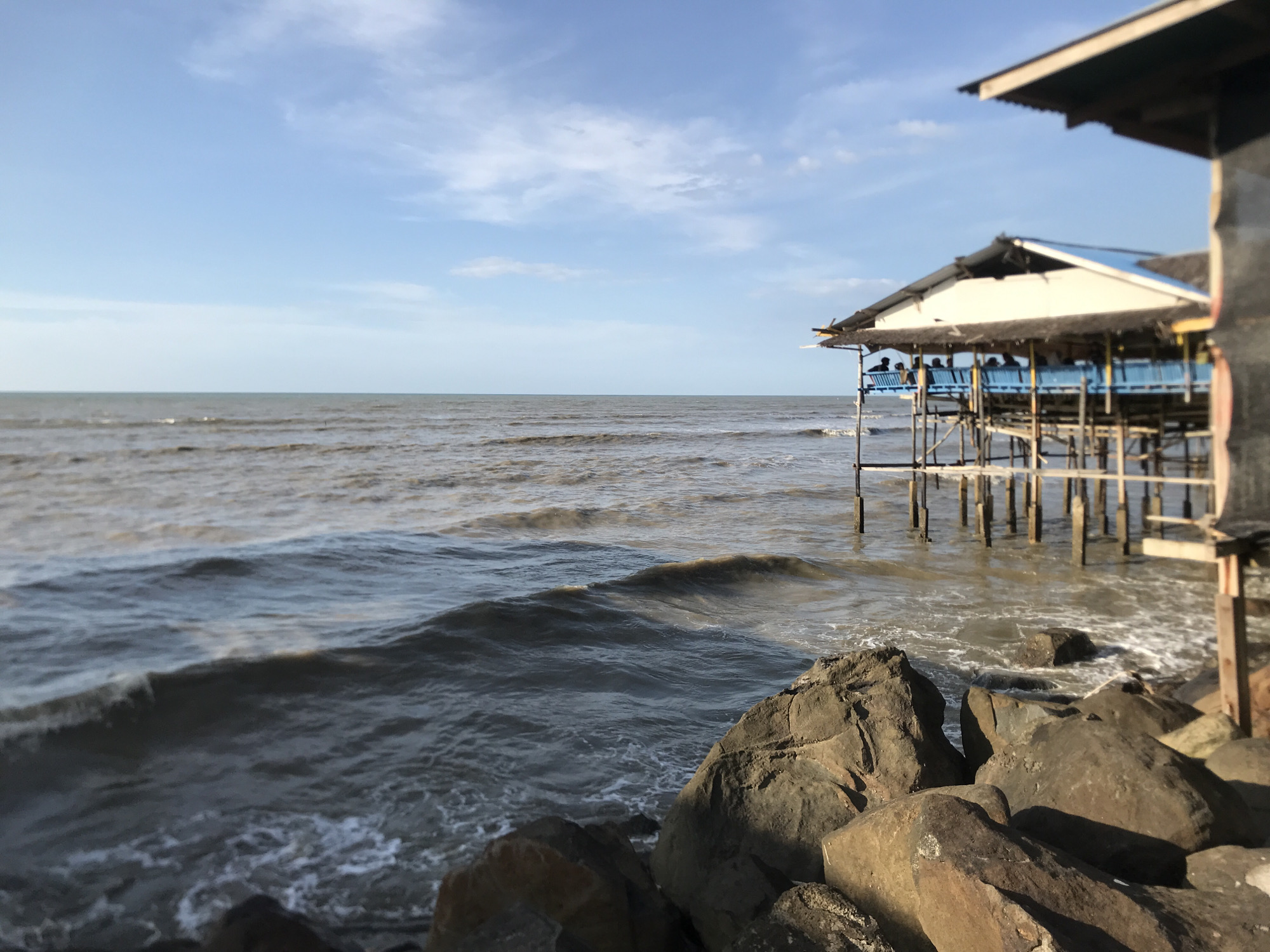 The image depicts a coastal scene in Lancok, Indonesia, featuring a stilt house extending into the sea. The house, constructed with wooden supports, has a white roof with a blue trim. The water is choppy, with small waves breaking against the rocky shoreline in the foreground. Several dark-colored rocks are piled along the shore. In the distance, the horizon line separates the sea from a partly cloudy sky. The structure's right edge is partially visible, suggesting the photographer's position is adjacent to another building. Several people are visible on the stilt house.