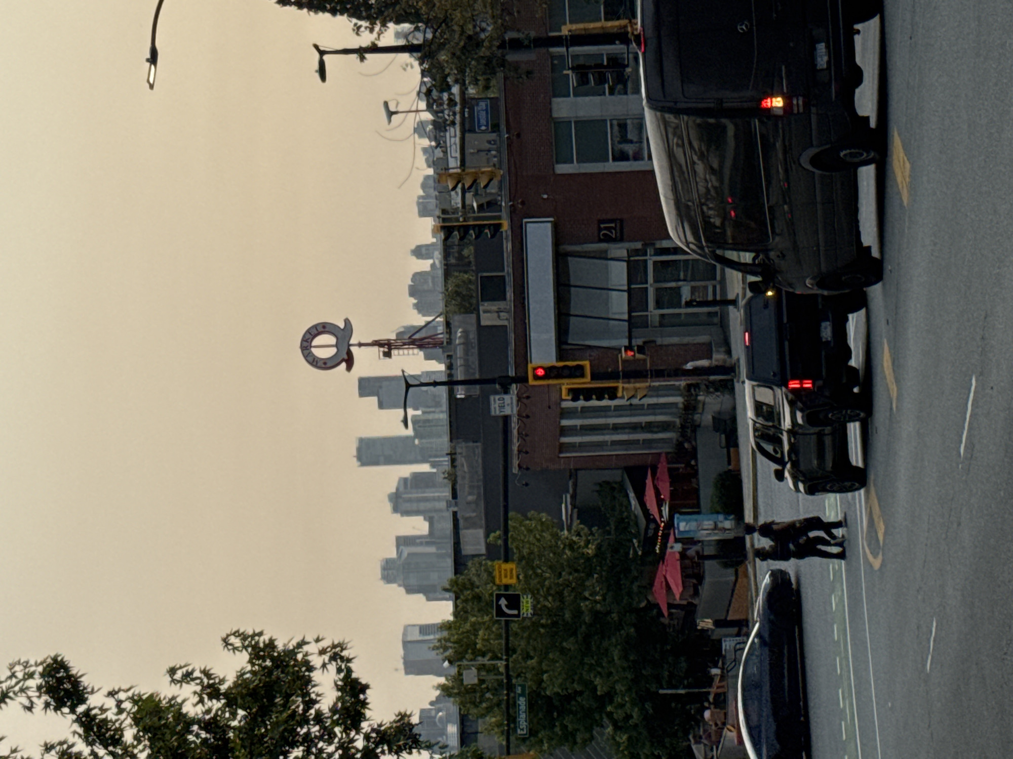 The image captures a street scene in North Vancouver, Canada, under a hazy, overcast sky. In the foreground, a black van and a black pickup truck are on the road, with the van's rear brake lights illuminated. Two pedestrians, dressed in dark clothing, are crossing the street in front of the pickup truck. To the left, a sleek black car is parked on the road.
The mid-ground reveals a brick building with large windows, identified by the number "21" on its facade. Red umbrellas are visible over outdoor seating areas at what appears to be a restaurant. Traffic lights, some illuminated red, are present. Road signs indicate "Esplanade" and a directional arrow with "NEW".
In the background, a cluster of modern skyscrapers forms a city skyline. A distinctive circular sign with "MARKET" is visible atop a structure. Trees frame the scene, with a large tree on the left and a smaller one on the right. Streetlights are also visible, one extending over the roadway. The overall impression is of an urban environment during daylight hours, possibly late afternoon given the subdued lighting.