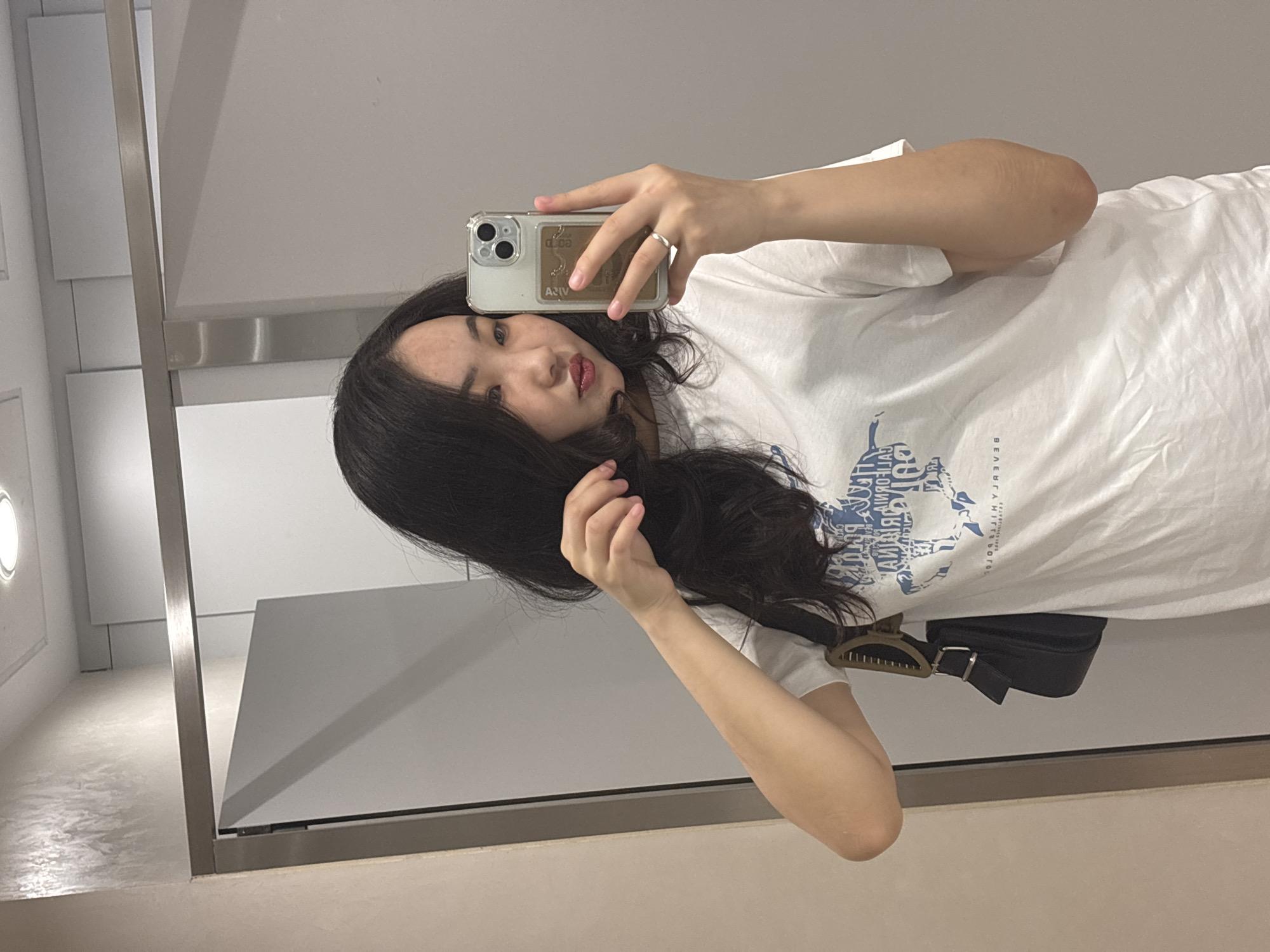 A young Asian woman with long, dark hair and dark eyes takes a mirror selfie in a public restroom. She is wearing a white t-shirt with a blue graphic design on the front. She is holding a silver smartphone in her right hand, and her left hand is touching her hair. A black purse with a silver buckle is slung over her left shoulder. The restroom has white walls and a metal stall door. The lighting suggests an indoor, possibly fluorescent, environment. The overall scene is mundane and personal, captured in a reflective surface. This type of scene is common in public restrooms found in various urban locations, including those in Astana, Kazakhstan.