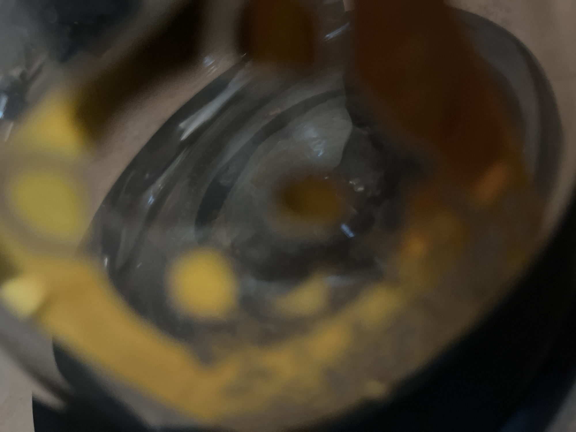 This is a close-up, slightly blurred photograph taken from above, looking down into a clear glass container. The container holds a swirling mixture of liquids, predominantly yellow and dark brown. The yellow appears to be a thick, possibly viscous substance, with streaks and patches visible. The dark brown liquid is translucent and seems to be mixing with the yellow, creating a marbled effect. There are reflections on the inner surface of the glass, suggesting it is clean.
The overall impression is that of a blending or mixing process taking place within the container. It's difficult to discern specific objects beyond the liquids and the glass itself, but the circular motion suggests a blender or a similar kitchen appliance is in use. The lighting is subdued, with some highlights on the glass, indicating artificial light rather than natural daylight. There are no discernible people, text, or environmental cues to determine the time of day, weather, or specific location. The focus is entirely on the contents of the container and the action of mixing.