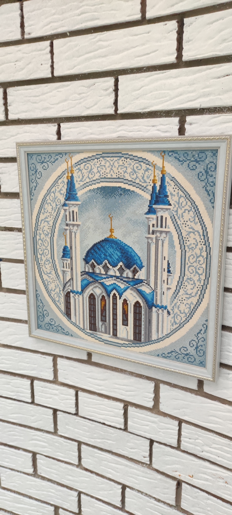 A framed cross-stitch depicting a mosque is mounted on a white brick wall. The mosque has blue domes and minarets with gold accents. The cross-stitch is enclosed within a circular, decorative border. The wall is made of rectangular, white bricks with visible dark grout lines. The image is taken from a slightly angled perspective, showing the texture of both the cross-stitch and the brickwork. There are no people or animals present in the image. The location in Rosinka, Russia is not visually discernible from the image.