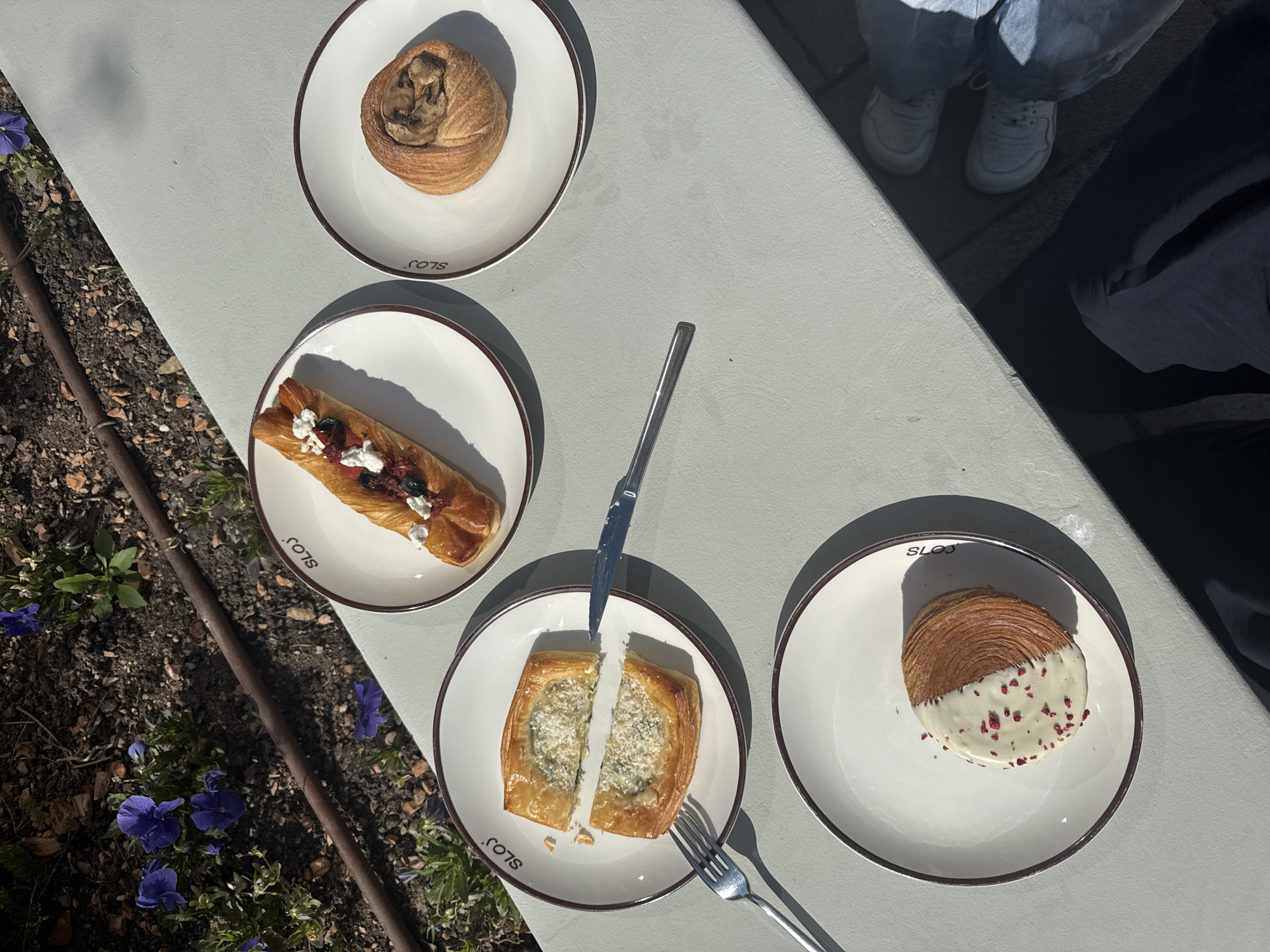 The image captures an outdoor dining scene on a sunny day, showcasing a light-colored table set with four plates of pastries. Strong shadows are cast, indicating bright sunlight.
Four white, round plates with dark brown rims are visible, each bearing the word "SLOJS" printed on its edge. On the top-most plate is a golden-brown, spiraled pastry, possibly a chocolate or nut roll, with a darker filling visible in its center. The plate to the middle-left holds an oblong puff pastry, generously topped with dark red fruit (likely berries), white creamy dollops, and dark, crumbled bits.
The bottom-middle plate features a square puff pastry that has been cut in half with a silver knife. The exposed interior reveals a savory green and white filling, possibly spinach and feta. A silver fork rests beside this plate. The fourth plate, located at the bottom-right, displays a round, layered pastry, similar to a cruffin or croissant swirl. One half of this pastry is covered in a smooth white glaze, speckled with small red flakes, possibly dried raspberries or rose petals.
The table is positioned next to a garden bed filled with dark soil, small stones, and patches of green plants. Several vibrant purple flowers, resembling pansies, are in bloom among the foliage. A dark, thin metal rod runs horizontally through the garden bed. In the upper right corner of the frame, part of a person's lower legs and feet are visible, clad in light blue jeans and white sneakers. A large, dark shadow of a person falls across the right side of the table and ground. The overall impression is one of a pleasant outdoor snack or meal.