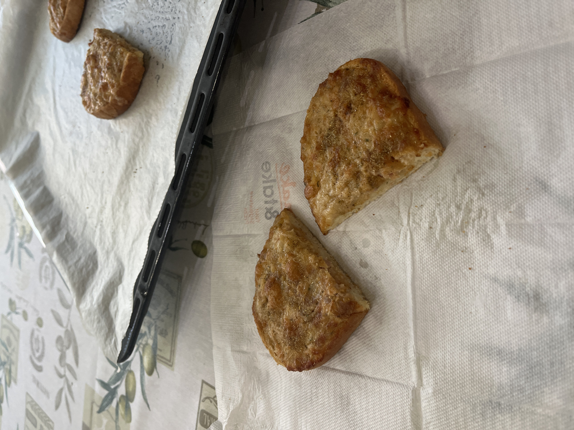 The image displays three golden-brown pieces of baked bread or pastry, likely garlic bread, resting on a white paper towel. Two pieces are prominent in the foreground, with another partially visible in the background. Each piece is irregularly shaped, appearing to be cut from a larger loaf, and is generously topped with visible herbs, possibly oregano or thyme, giving it a seasoned appearance.
Beneath the baked items, a crinkled white paper towel with a distinct textured pattern is laid out, presumably to absorb excess oil or moisture. A black metal baking tray or oven rack runs along the left side of the image, suggesting the items have recently been removed from it. Underneath the paper towel, a decorative tablecloth with a light background is partially visible, adorned with a pattern of green olive branches and olives.
The scene depicts food preparation or serving in a domestic setting, with the baked goods cooling after being cooked. The lighting suggests an indoor environment. The photograph is taken from a slightly elevated, angled perspective.
Visible text includes the word "Blake," which is printed on the paper towel, appearing rotated and partially obscured. The context indicates this scene takes place in Sofia, Bulgaria, although no specific visual elements within the image directly confirm this geographical location.