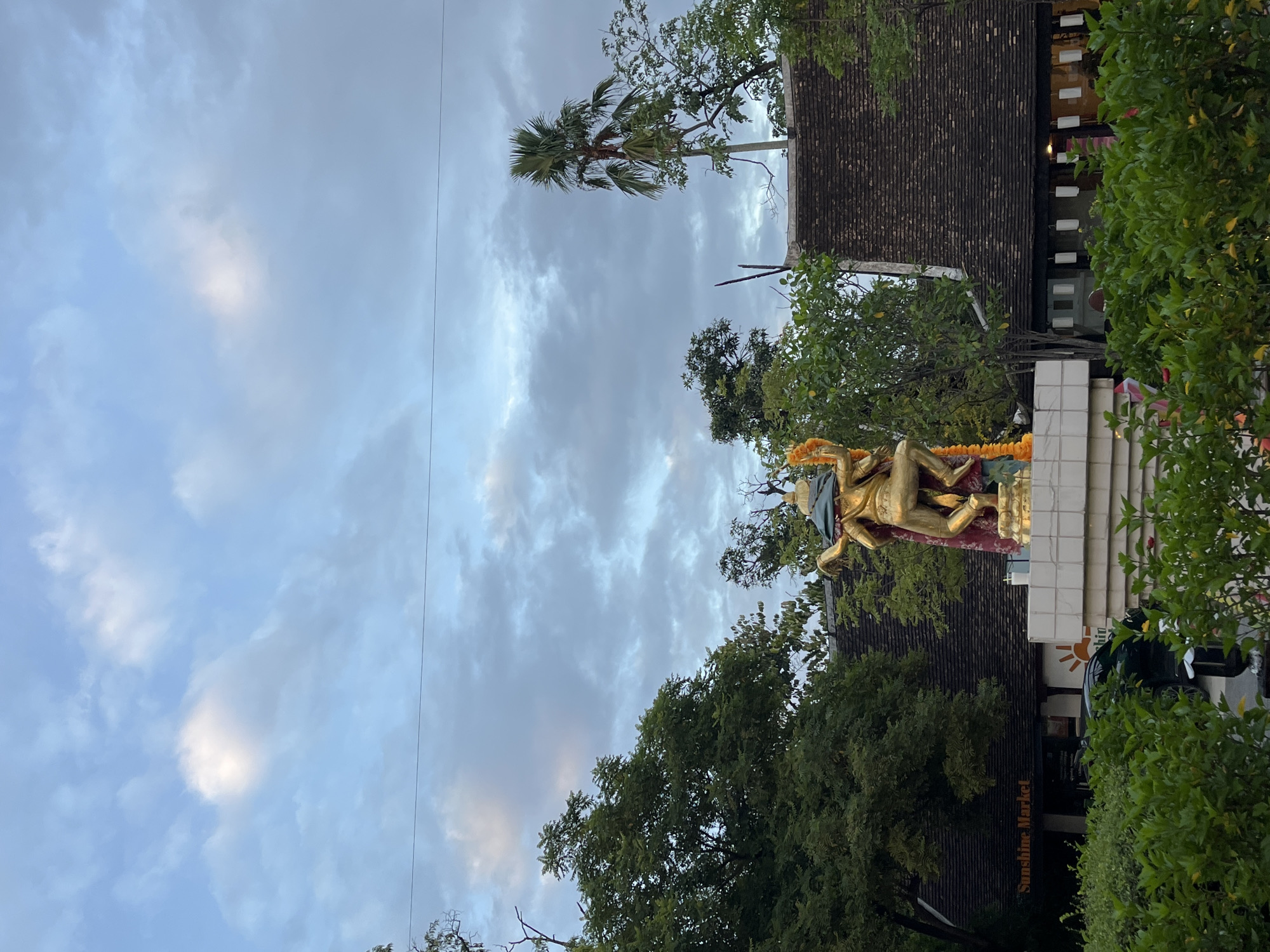 An outdoor, eye-level medium shot captures a golden statue of a deity with multiple arms in a dynamic pose, adorned with a vibrant orange marigold garland. The statue stands on a white pedestal in front of buildings with dark, tiled roofs. Lush green trees and bushes surround the area, with a tall palm tree visible to the right. The sky above is a soft blue, filled with wispy clouds, suggesting it is either early morning or late afternoon. A sign on the left building reads "Sunshine Market." The scene is likely in Chiang Mai City Municipality, Thailand, given the architectural style and tropical vegetation. There are no people clearly visible in the shot.